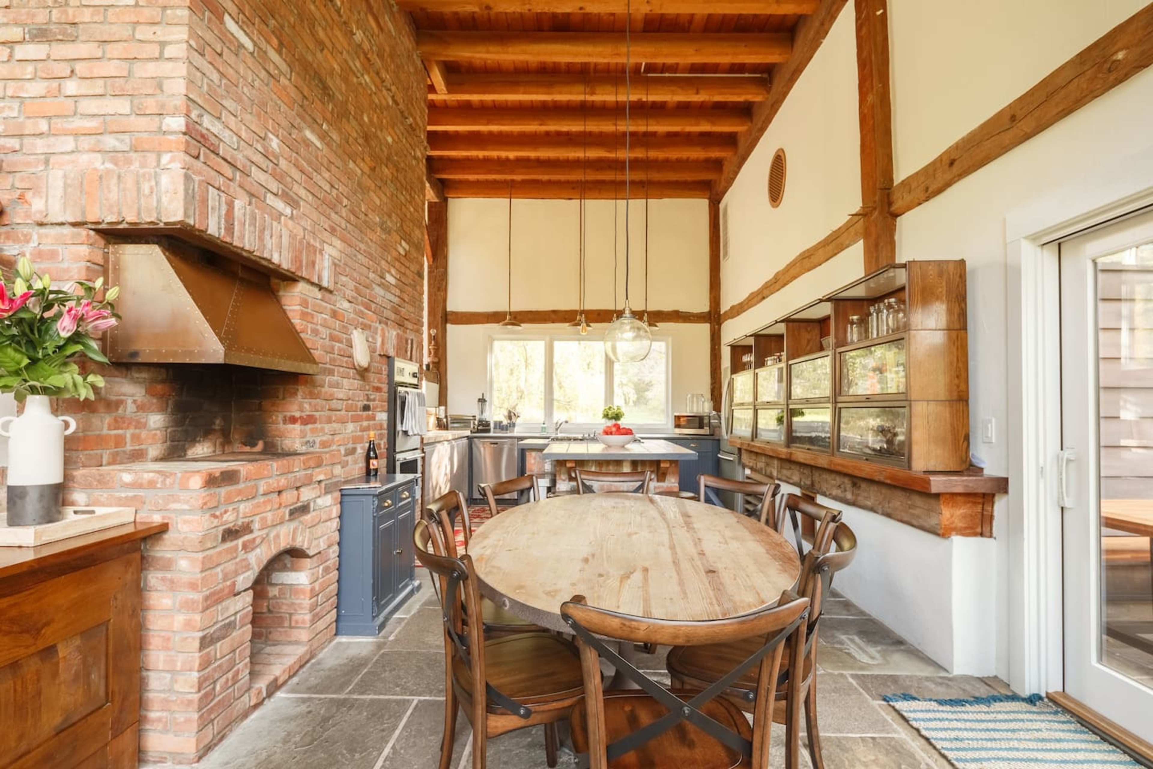 A spacious kitchen with a large wooden dining table, exposed brick walls, a high ceiling with wooden beams, and a modern kitchen setup in the background.