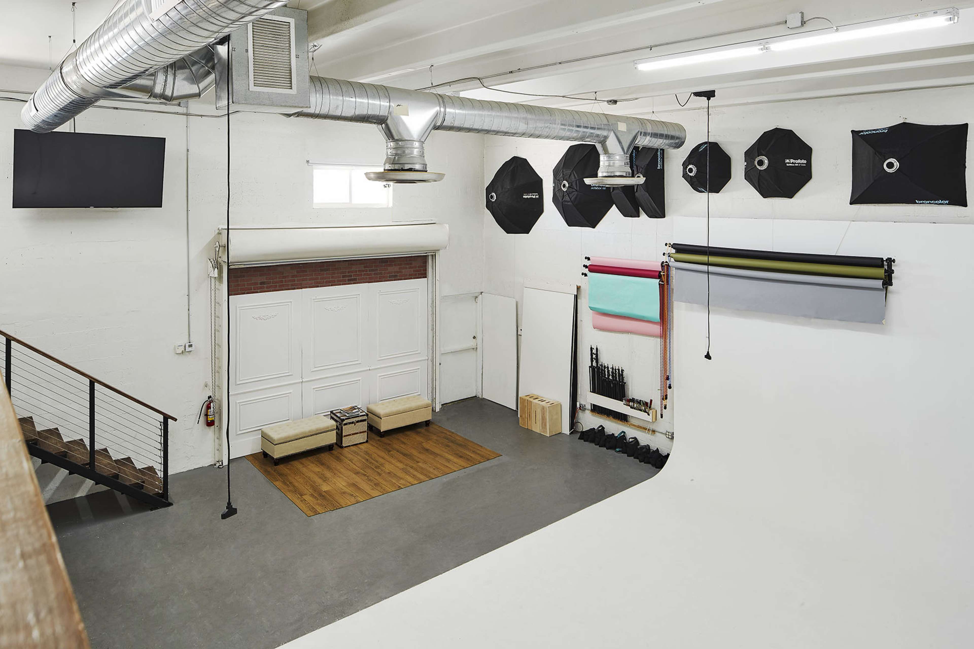 The image shows a spacious photography studio featuring a white backdrop, various lighting equipment, and a seating area with a wooden floor.