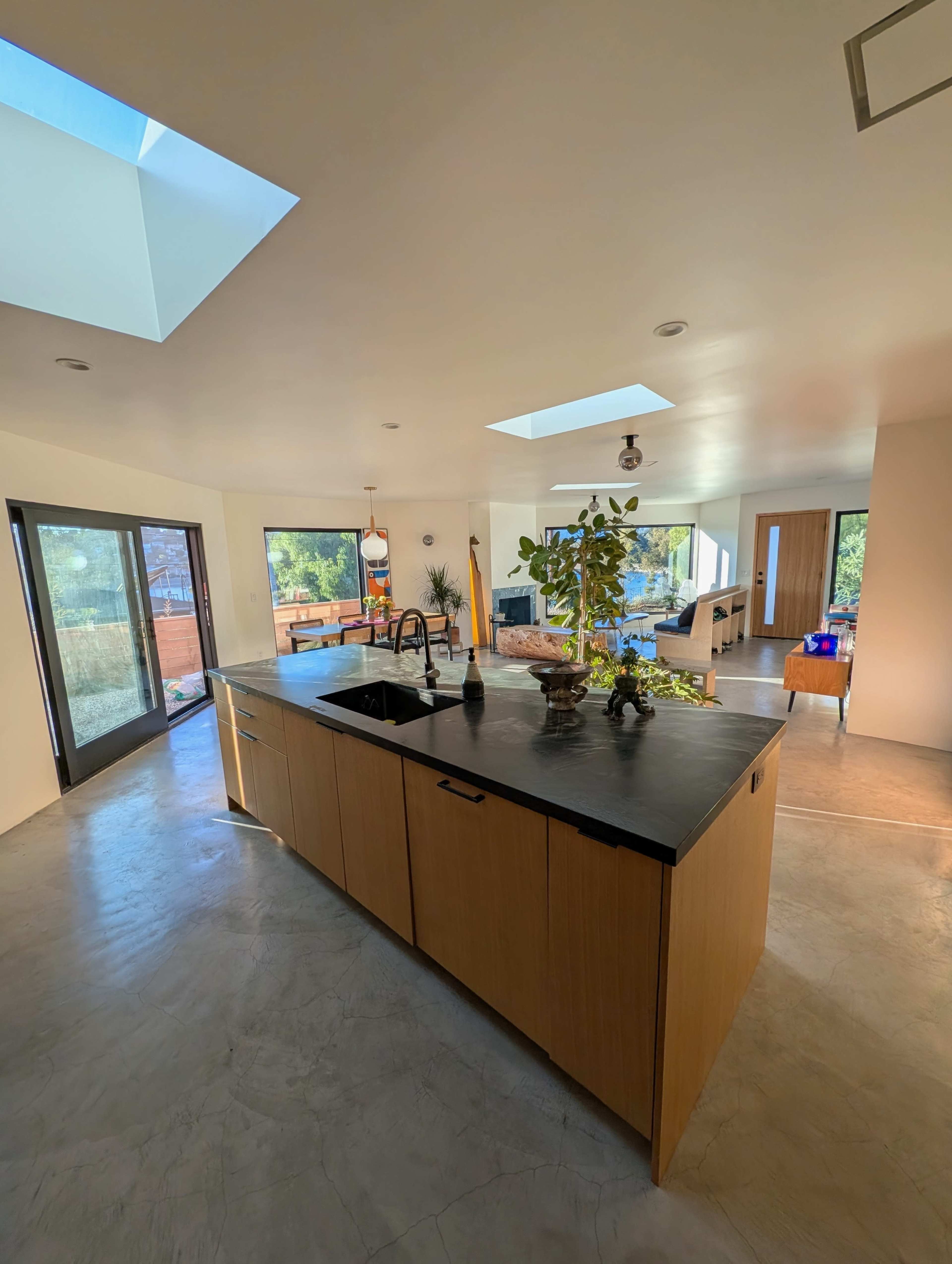 A modern kitchen features a central island with a dark countertop, surrounded by large windows and skylights that illuminate the open space.