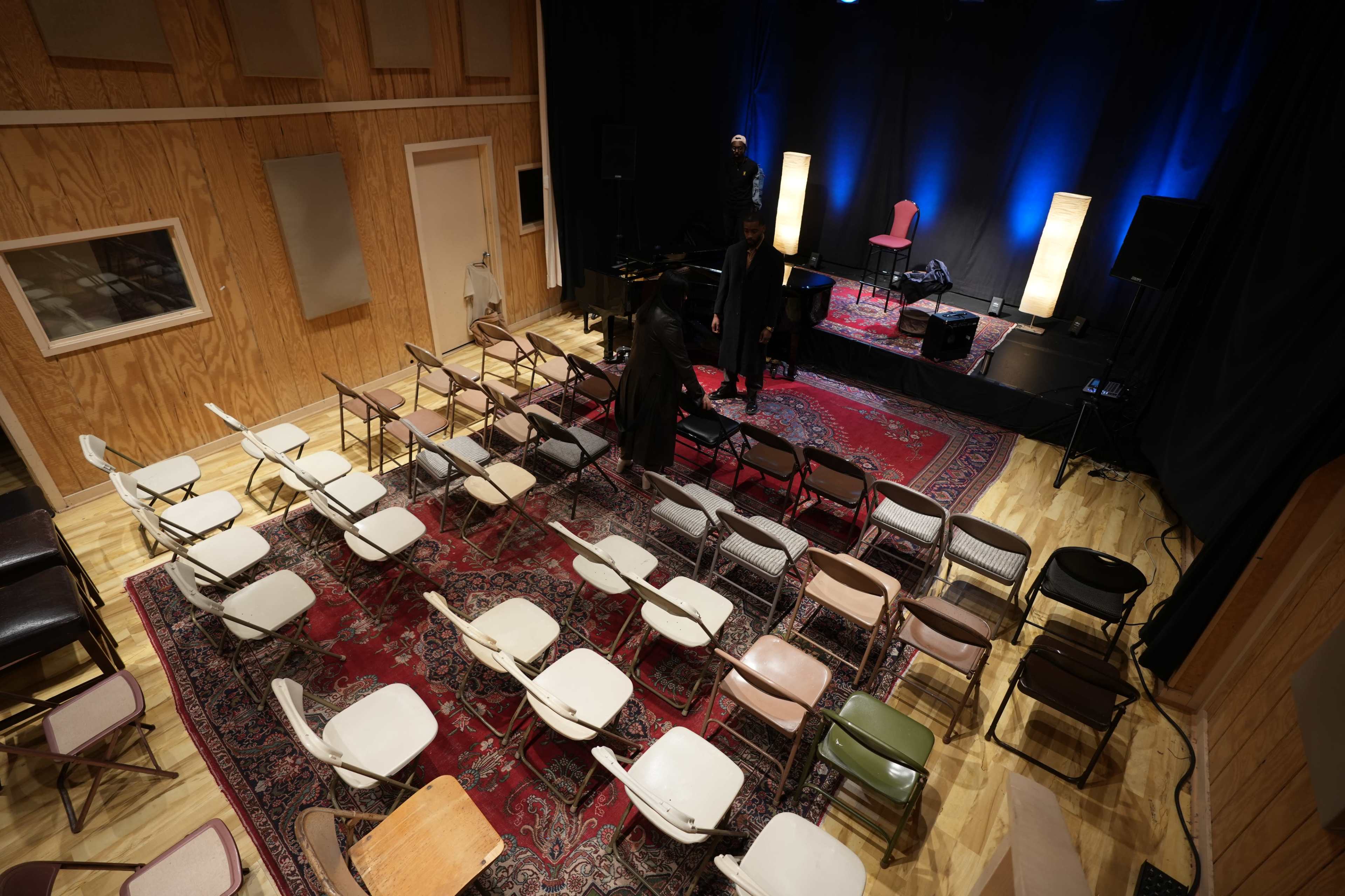 A small performance space with rows of chairs arranged on a wooden floor, a small stage set up in the background with blue lighting, and a few people preparing on stage.