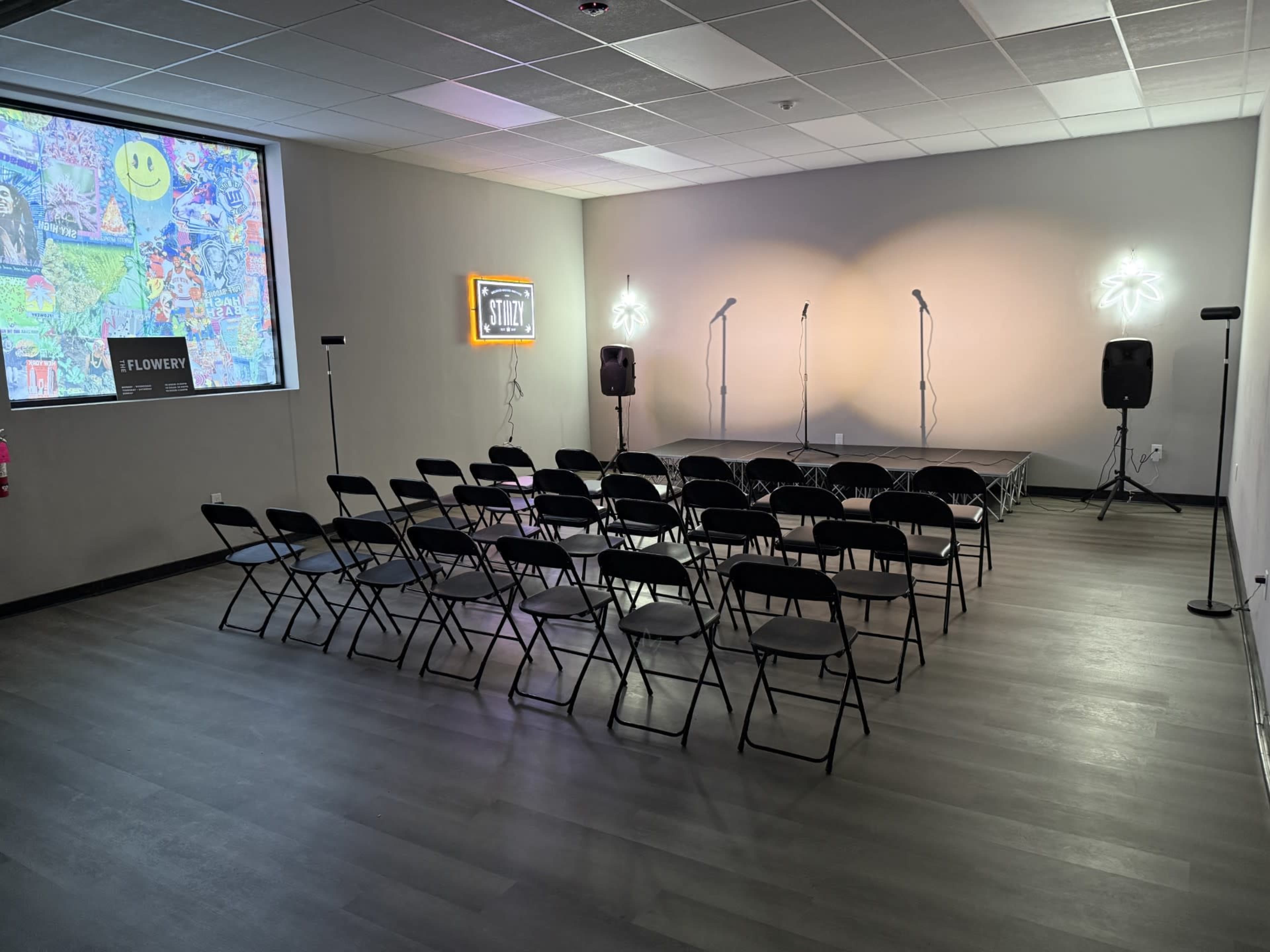 The image shows a small stage with three microphones, surrounded by rows of black folding chairs, and a colorful mural on one wall.