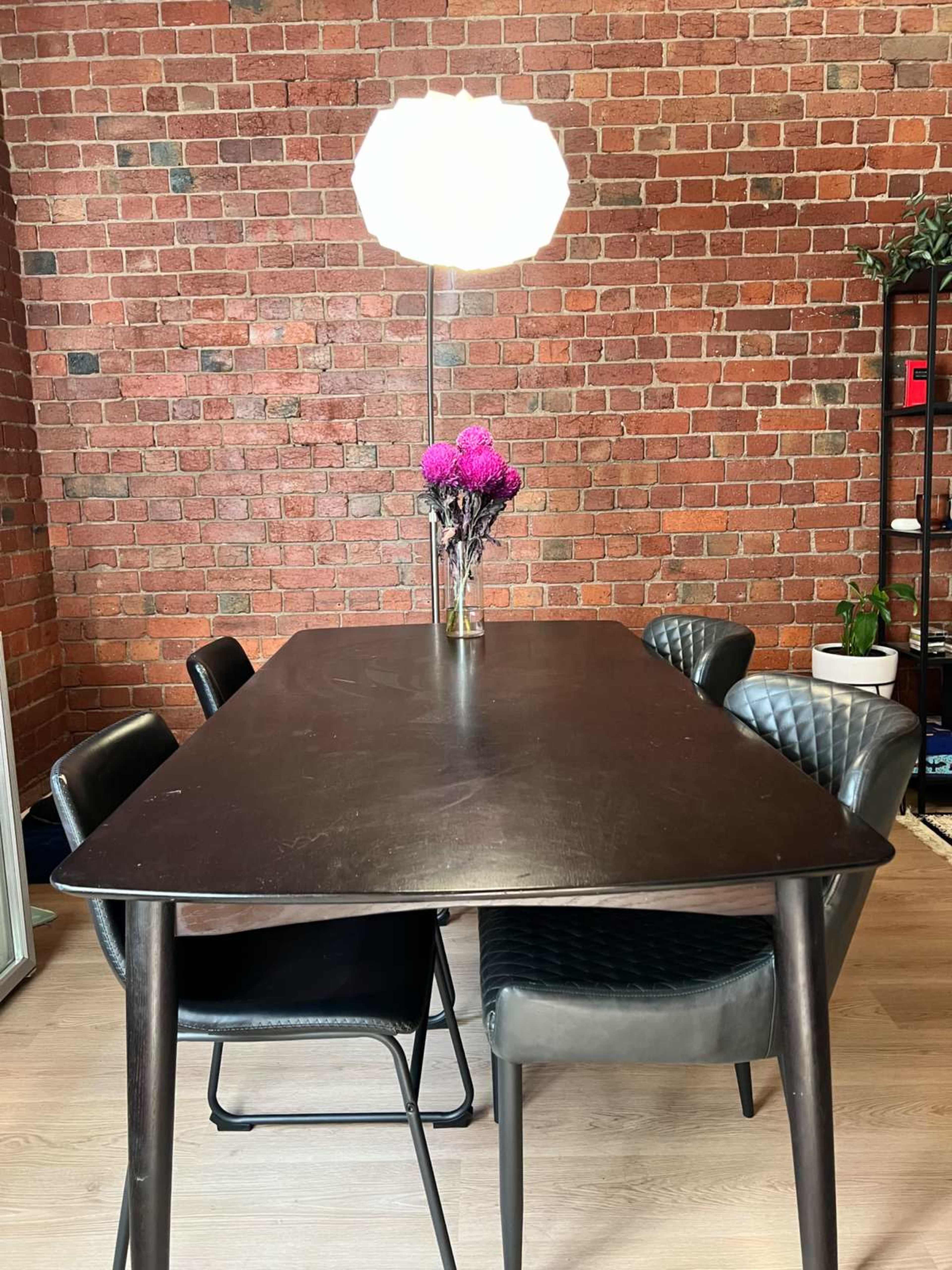 A dark wooden table with four chairs and a vase of flowers is set against a brick wall, illuminated by a modern pendant lamp.