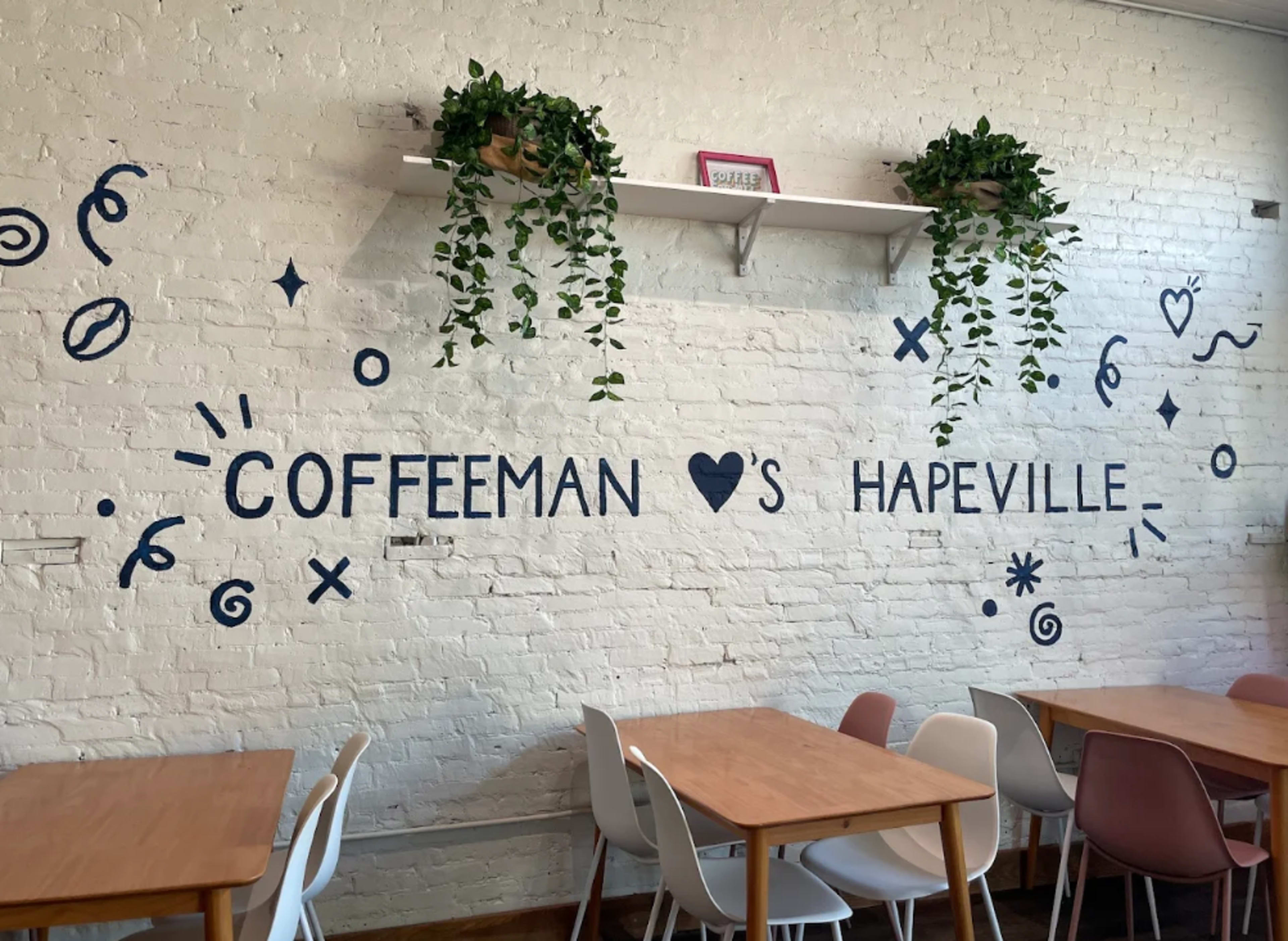 The image features a white brick wall adorned with green plants and blue decorative designs, displaying the text "COFFEEMAN LOVES HAPEVILLE" above wooden tables and chairs.