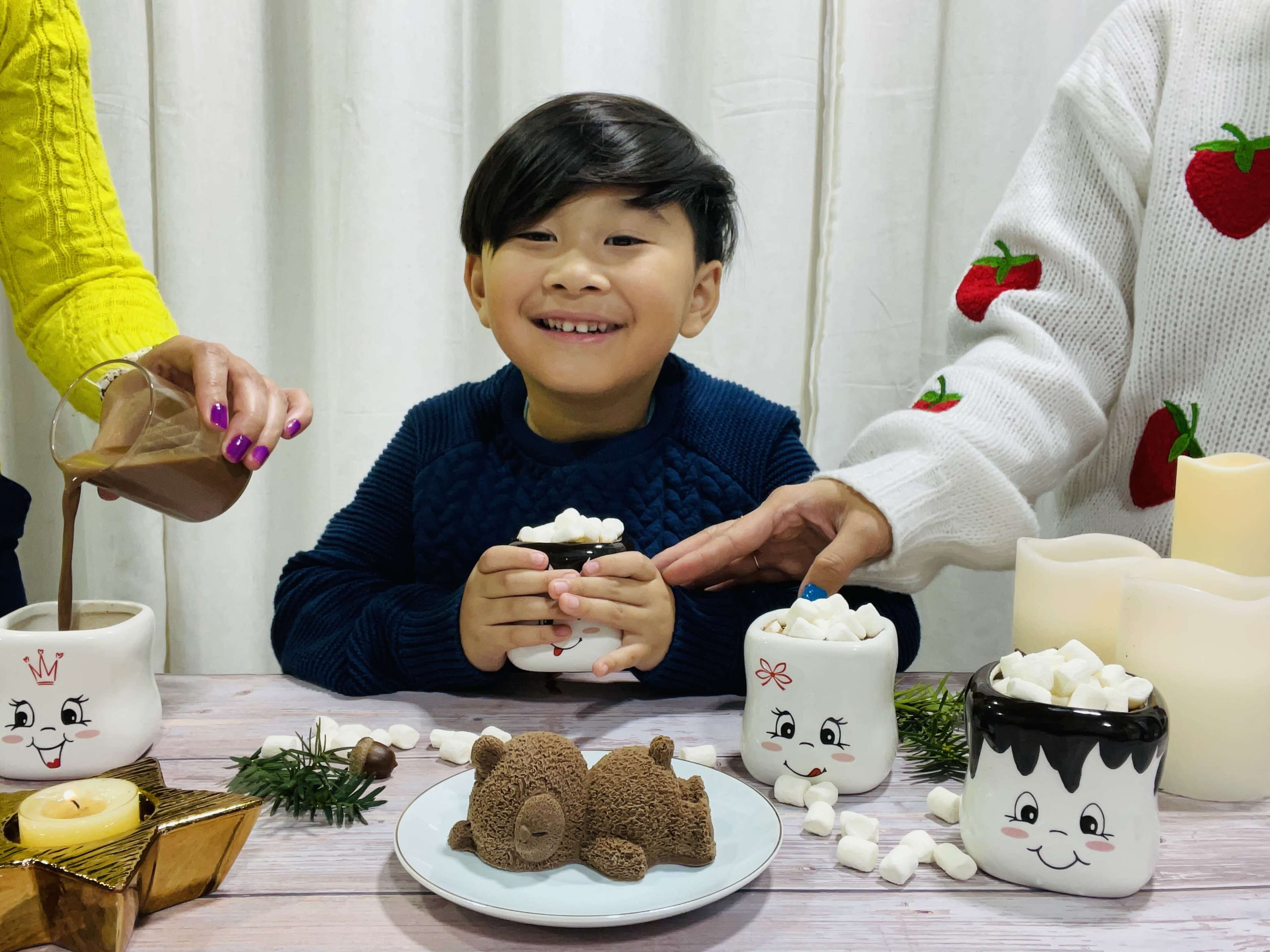 A boy smiles while holding a mug filled with marshmallows, surrounded by candles and a chocolate bear dessert on a table.