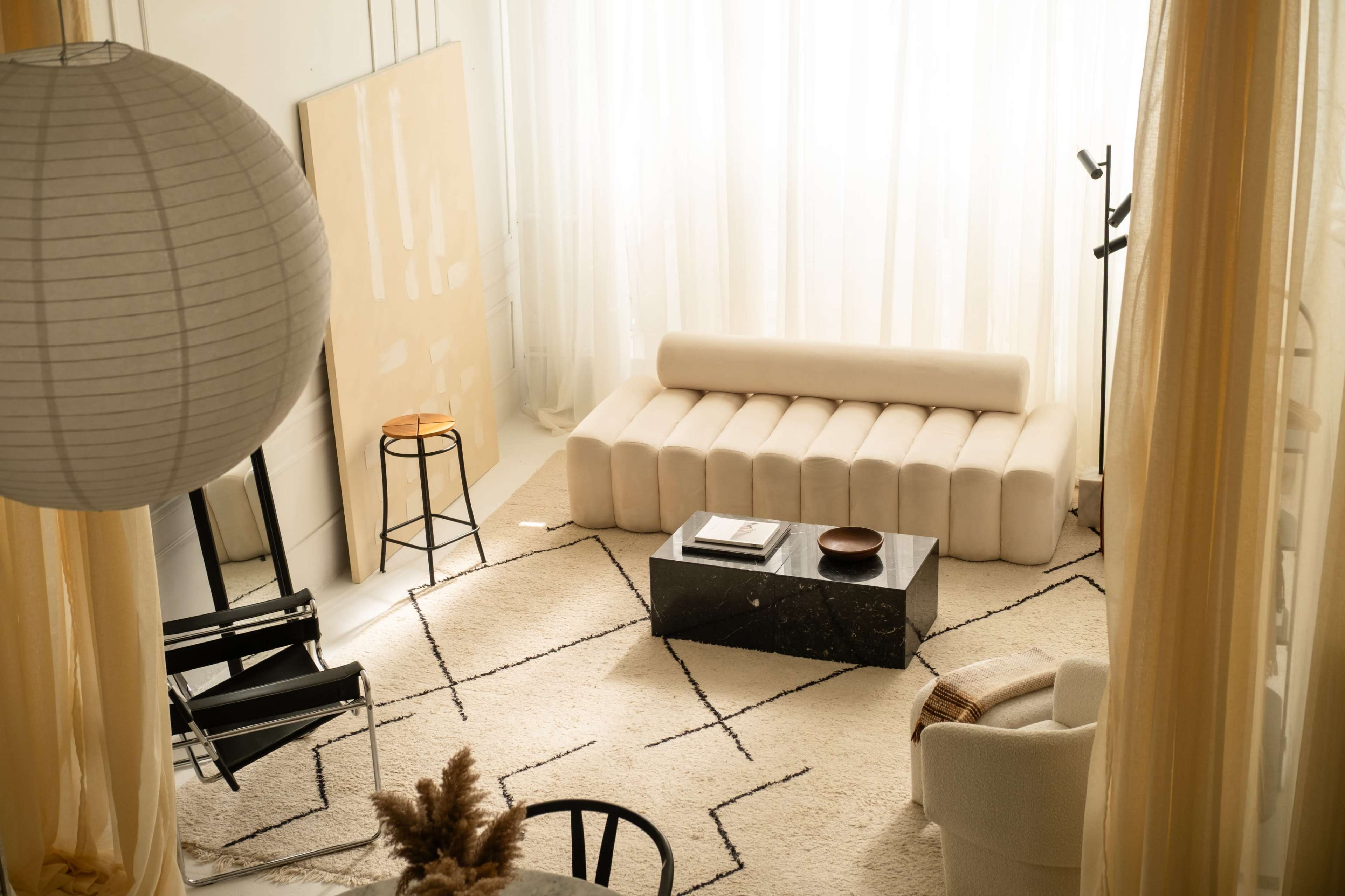 The image shows a modern, minimalist living room with a beige sofa, a black coffee table, and large windows covered by sheer curtains.