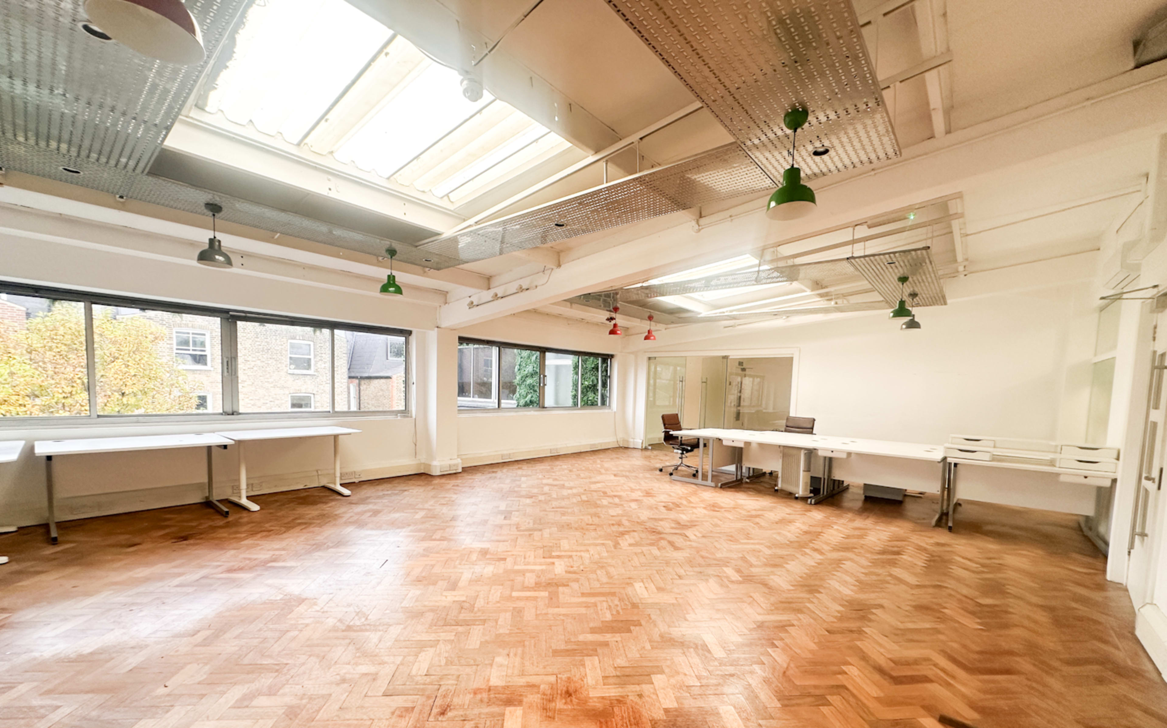 The image shows an empty office space with large windows, wooden herringbone flooring, and several workstations.