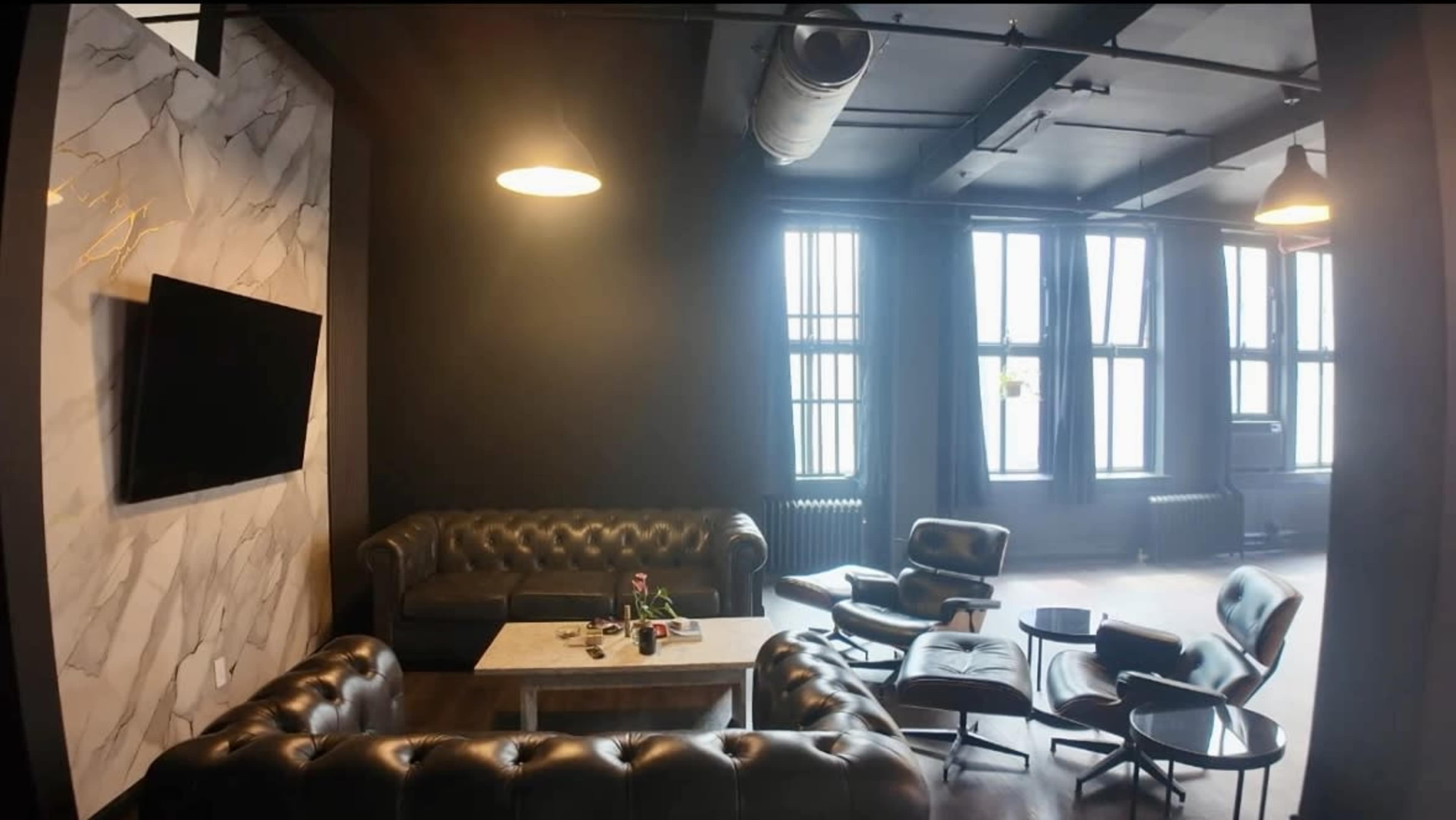 The image shows a modern lounge area with two leather sofas, a coffee table, and several armchairs, illuminated by overhead lighting and natural light from large windows.