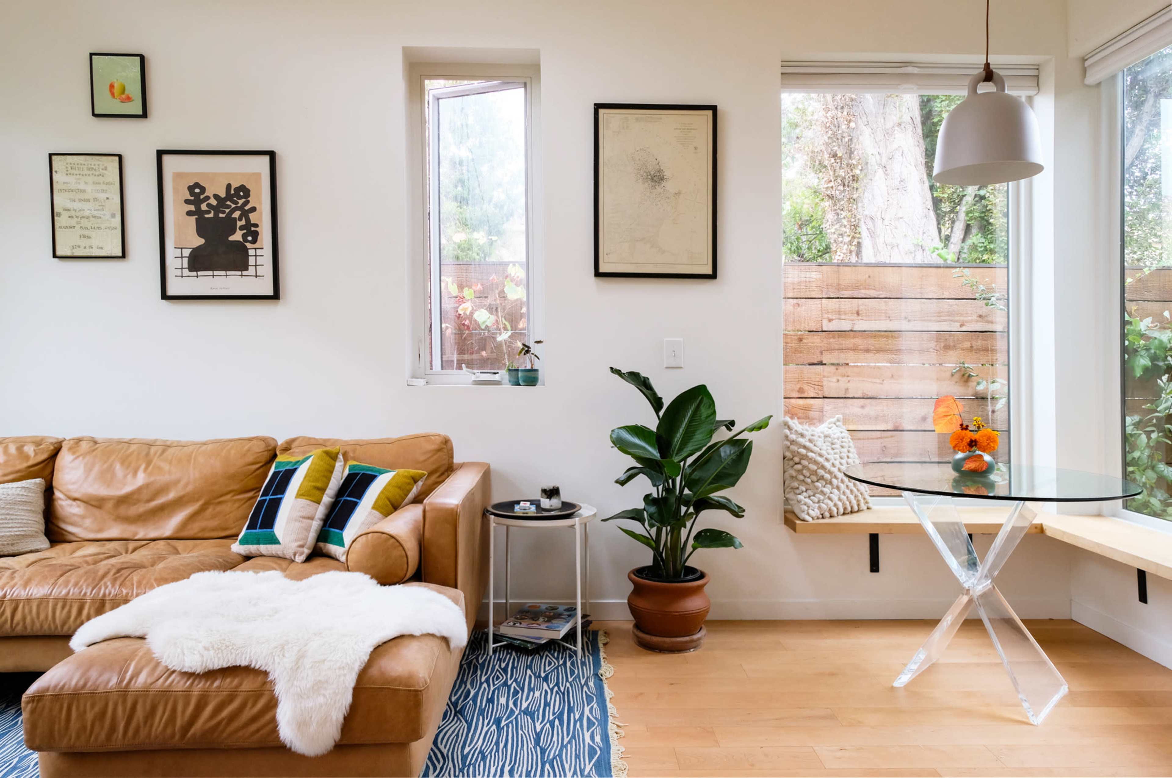 A modern living room features a leather couch, a glass dining table, and large windows with plants and a wooden fence outside.