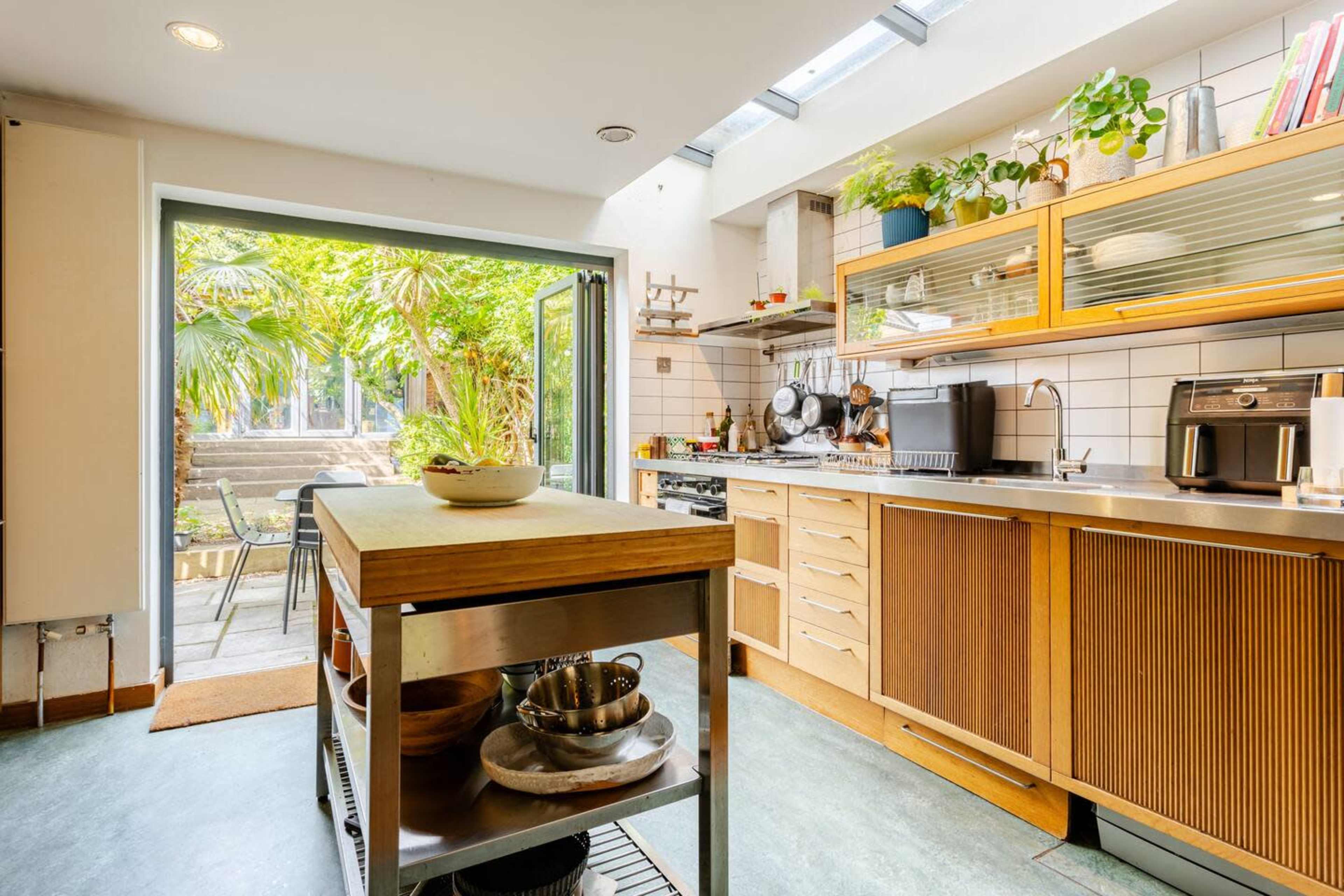 The image shows a modern kitchen with wooden cabinets and an island, featuring an open door that leads to a garden with greenery visible outside.