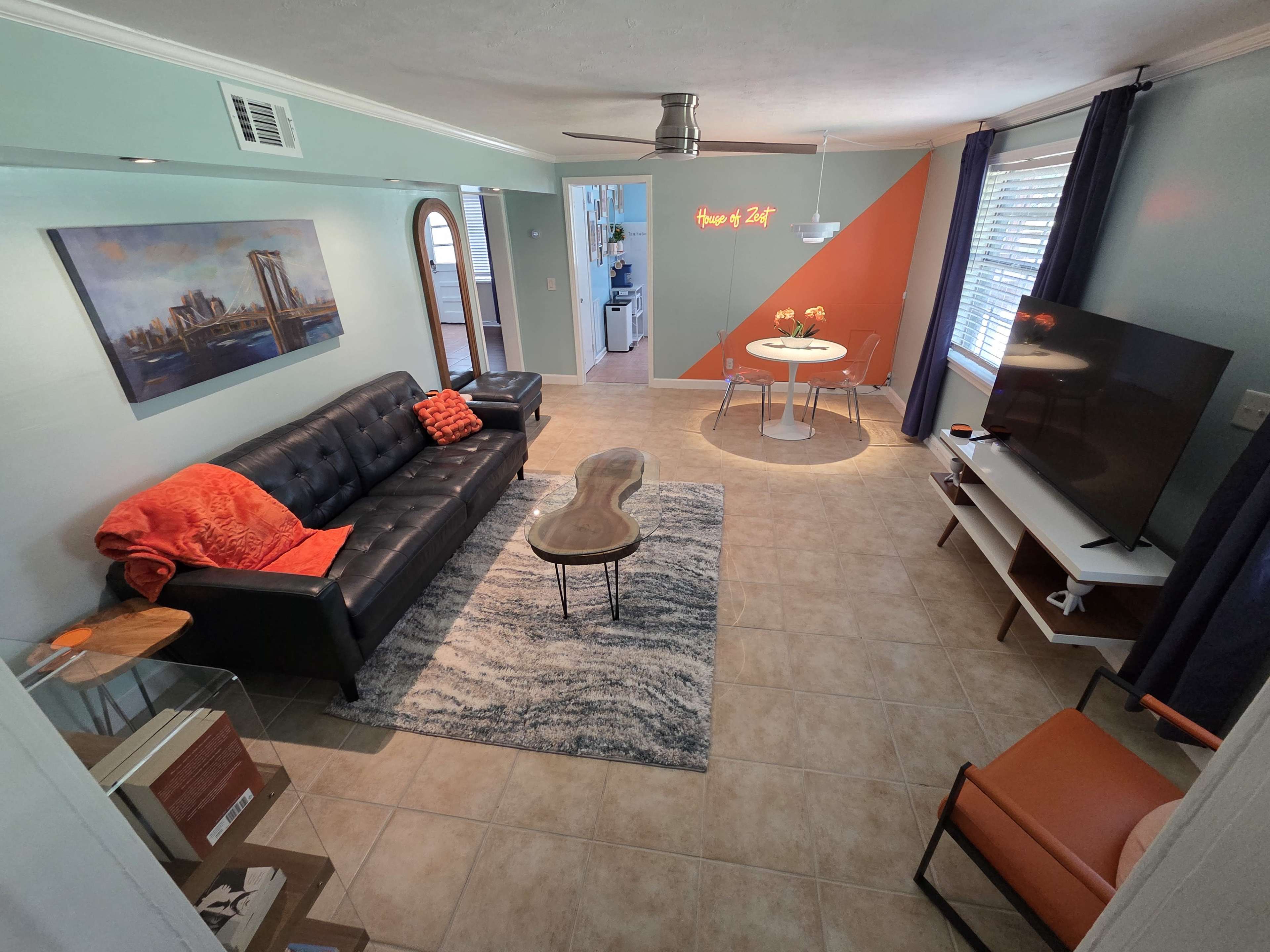 The living room features a black leather sofa, a wooden coffee table, and a circular dining table with chairs, accented by an orange wall and decorative lighting.