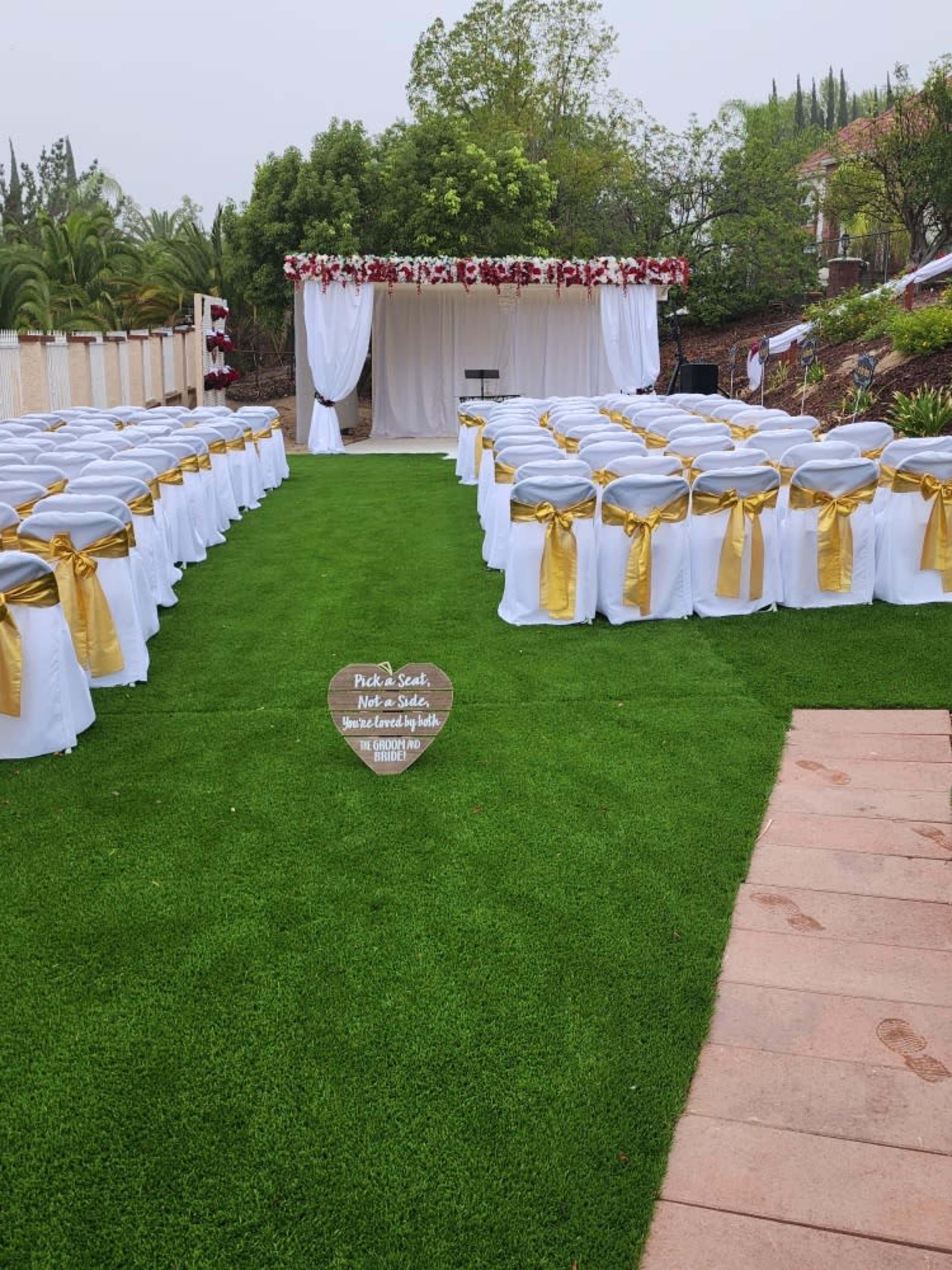 A wedding ceremony setup features rows of white chairs adorned with gold ribbons facing a decorated altar under a canopy.