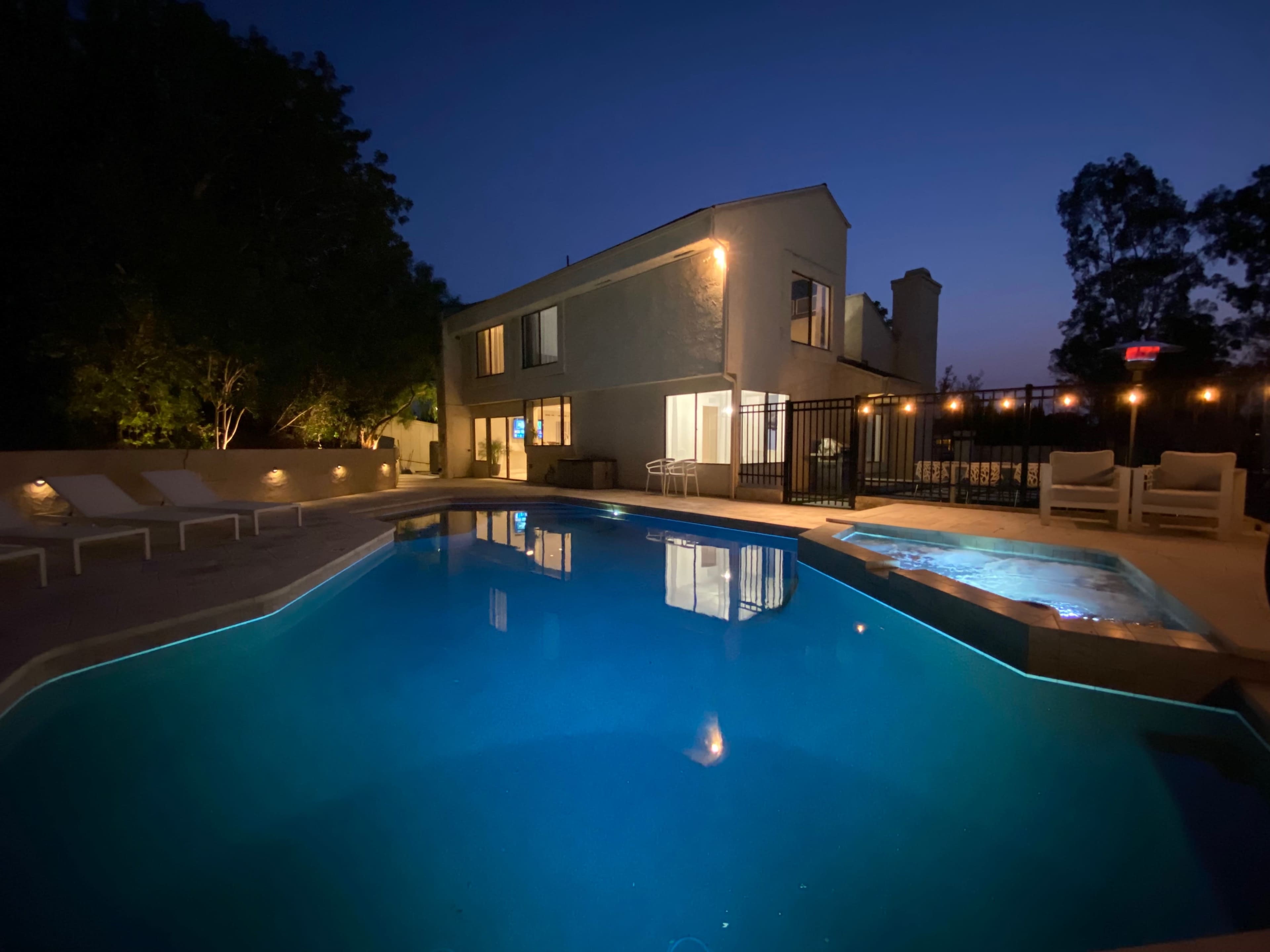A house with a pool and lounge chairs is illuminated at night, surrounded by trees and a fence.