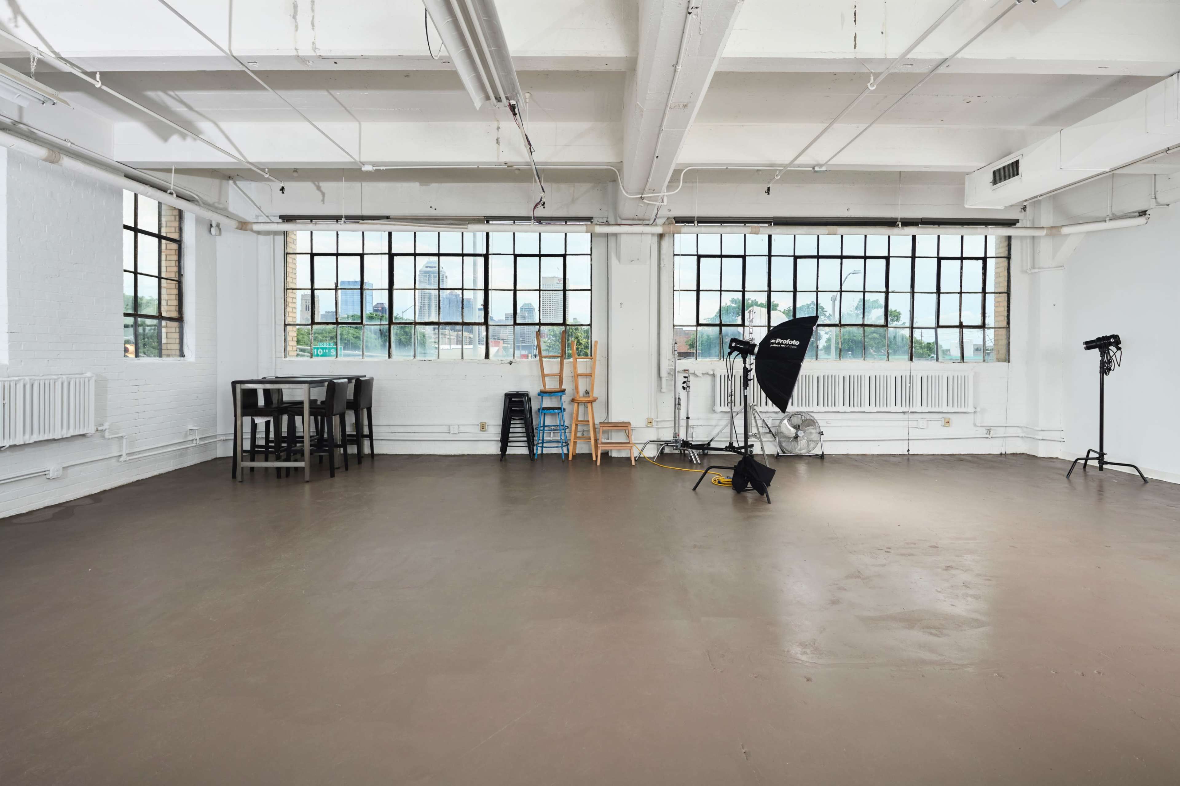 The image shows a spacious, industrial-style studio with large windows, a bare concrete floor, a table and chairs, and photography equipment in one corner.