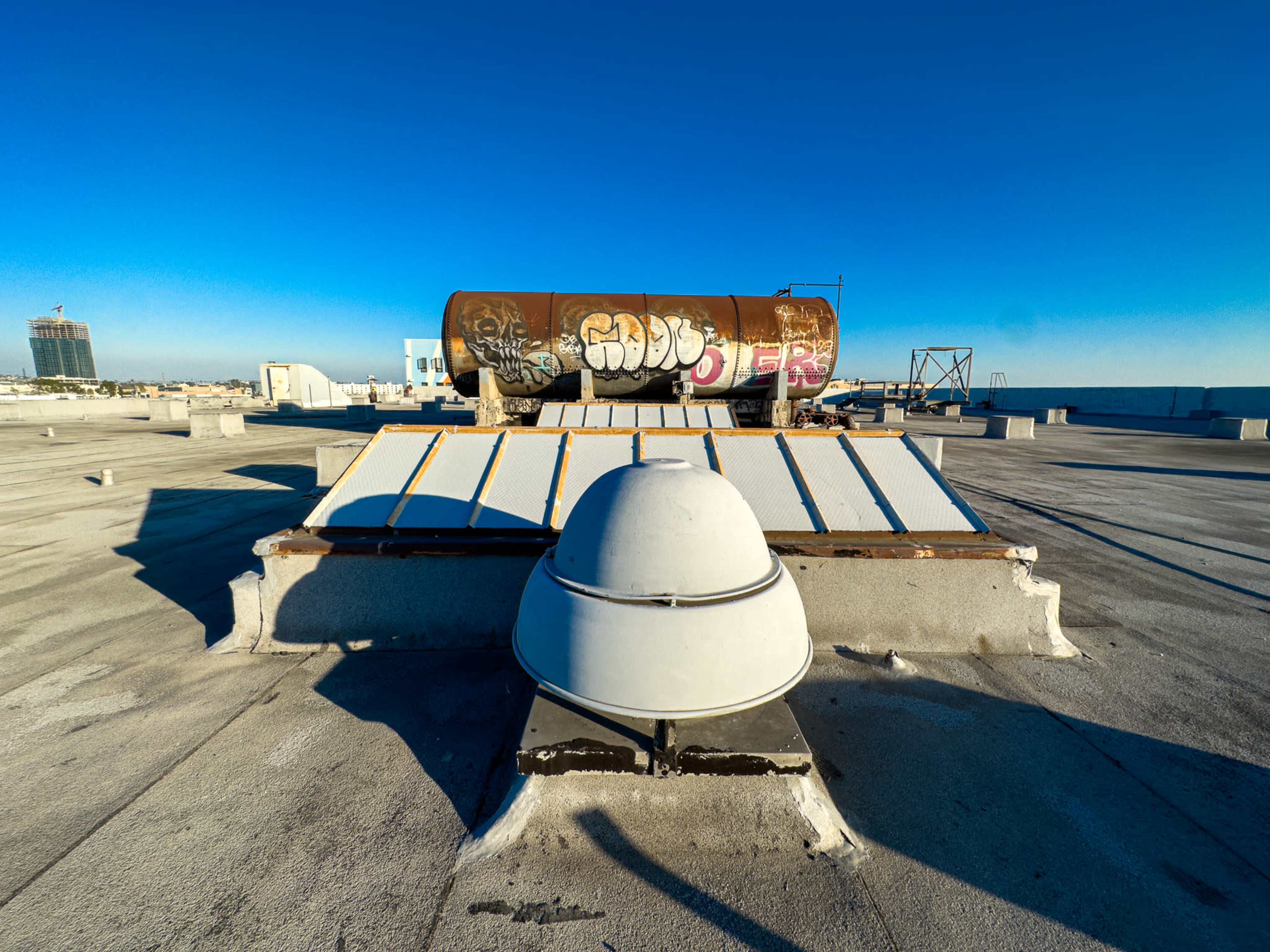 A rooftop scene featuring a large, rusty tank with graffiti and a white ventilation structure in the foreground against a clear blue sky.