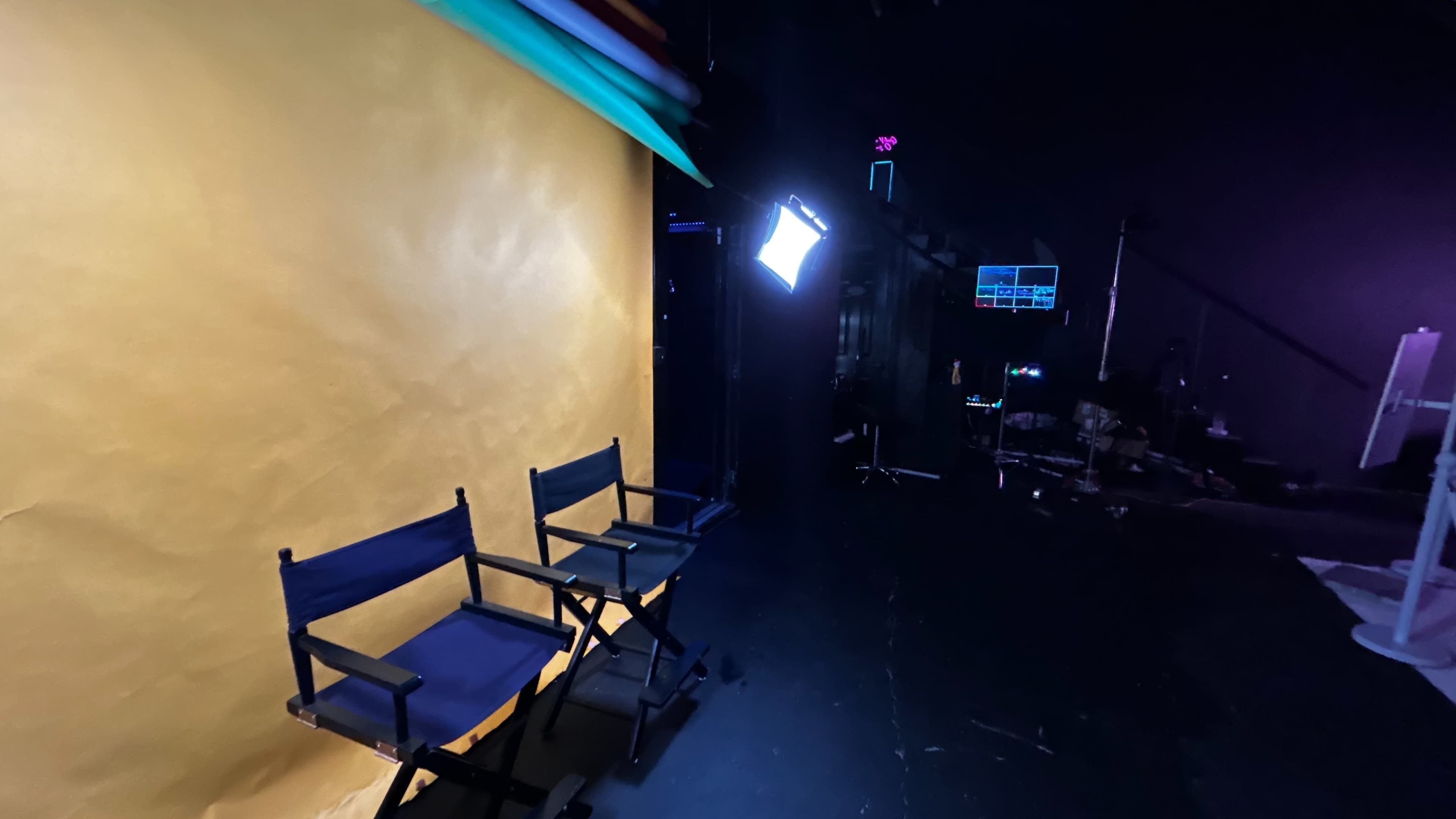 The image shows two director's chairs in front of a yellow backdrop with studio lighting and equipment in the background.