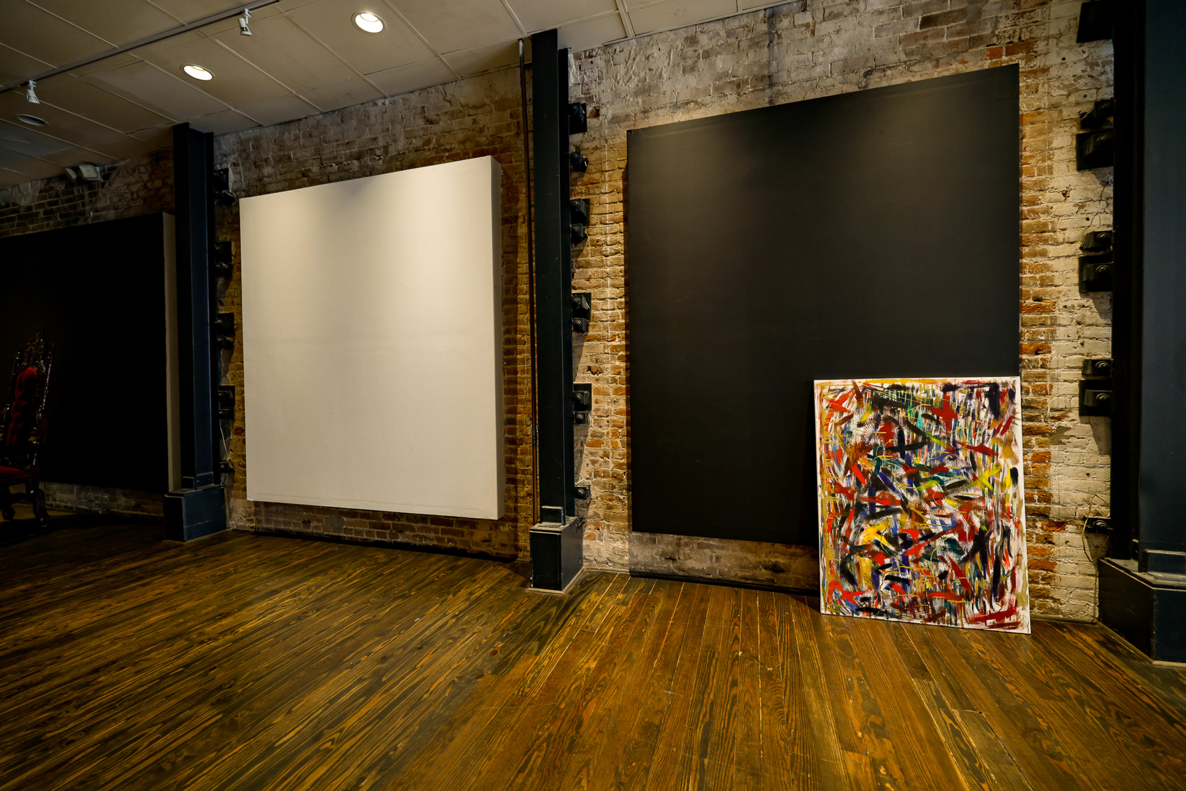 An art gallery interior with exposed brick walls, featuring two large blank canvases and one colorful painting on the floor.