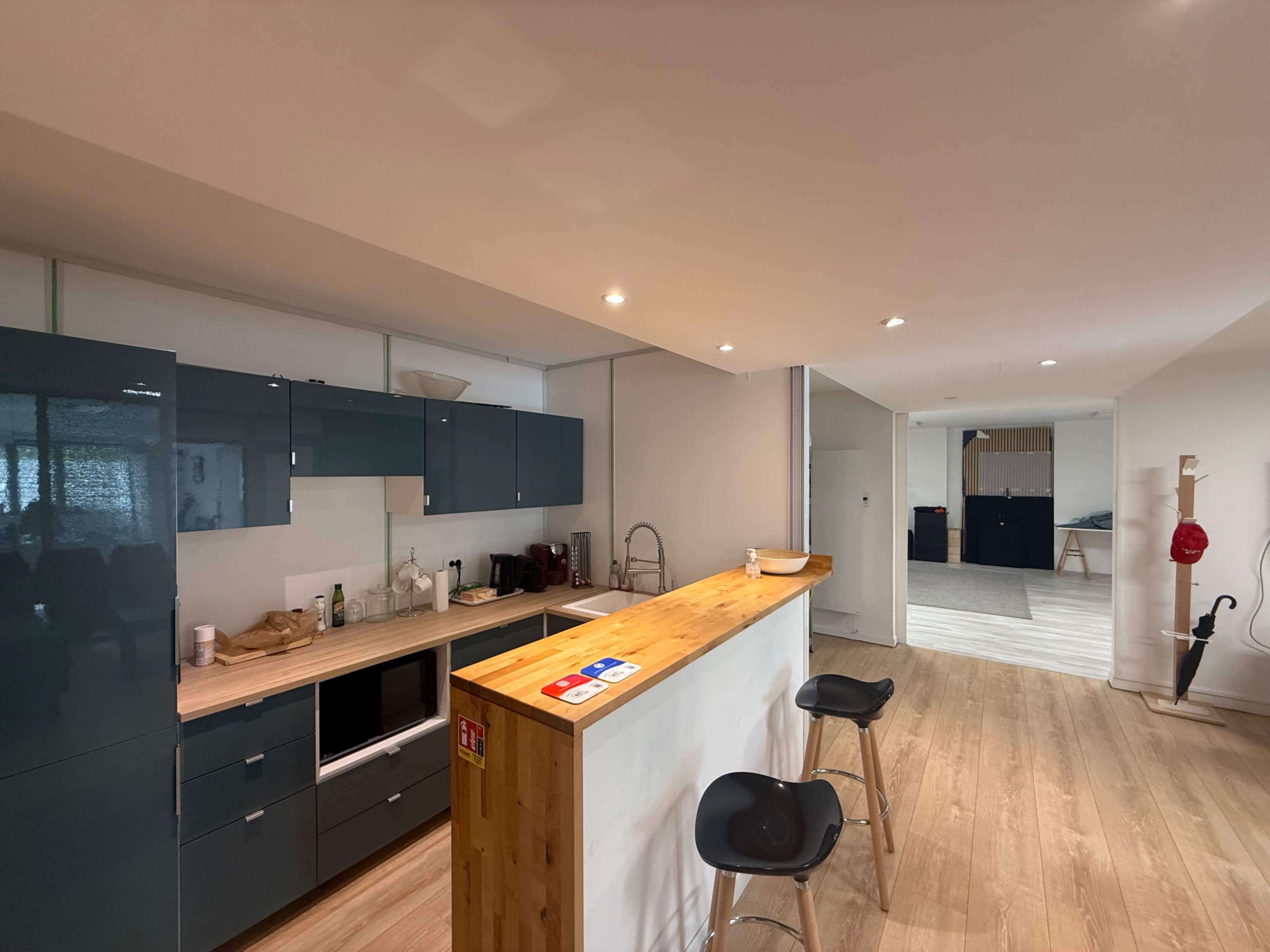 A modern kitchen features sleek cabinets, a wooden countertop, and two bar stools, with an open layout leading to an adjacent room.