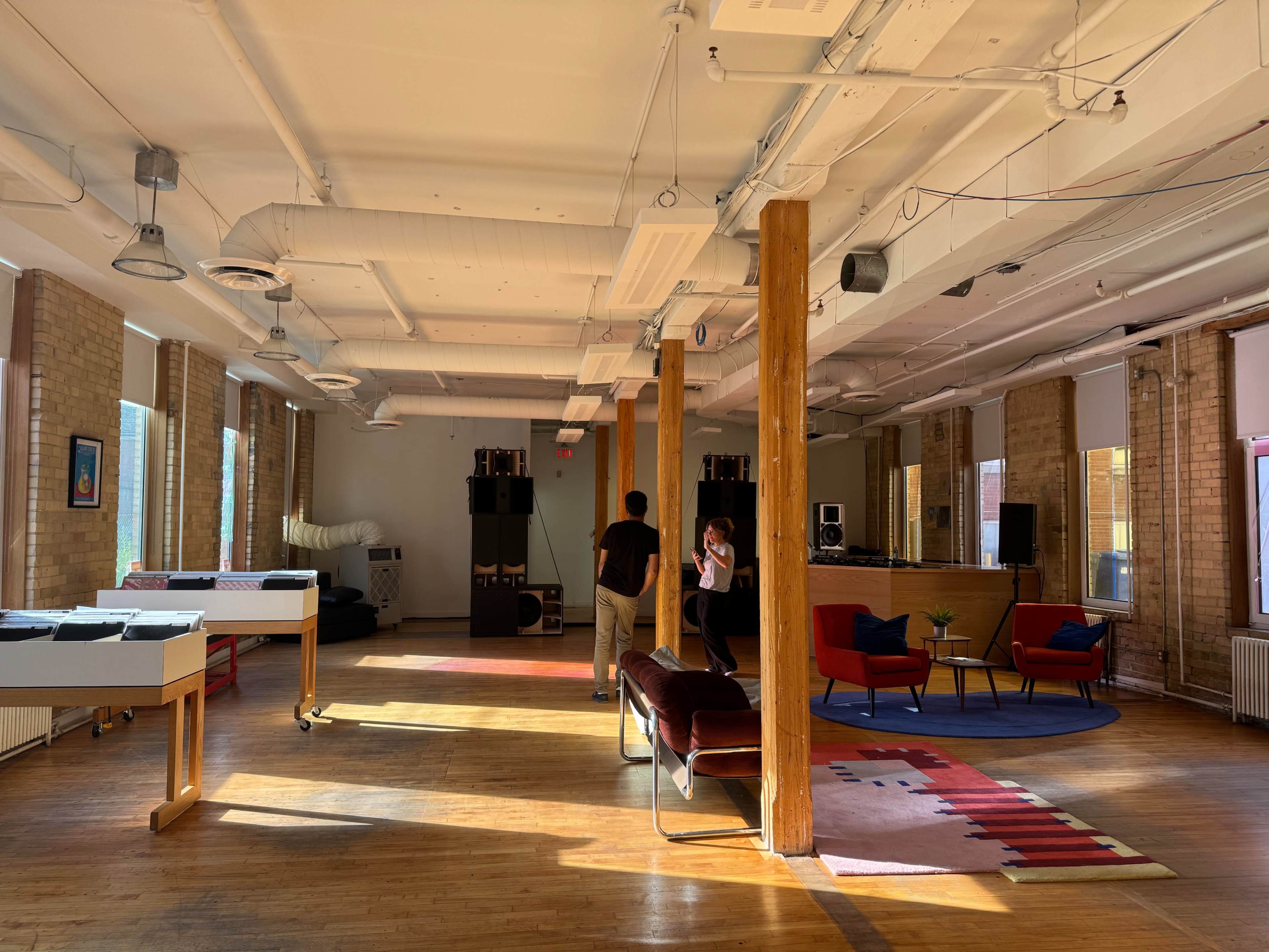 The image shows a spacious, well-lit room with wooden floors, featuring a few pieces of furniture including chairs, a couch, and a game table.