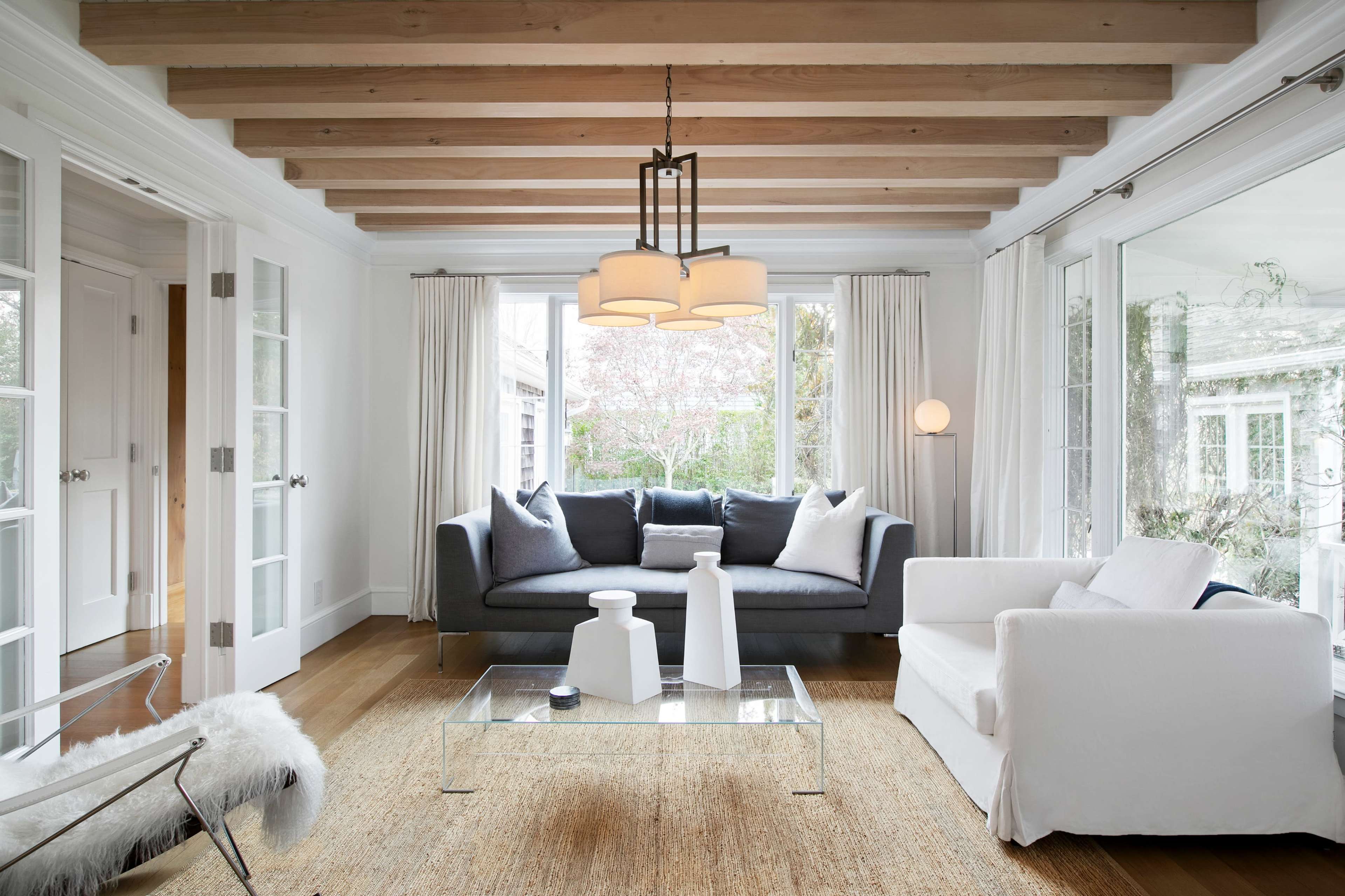 The living room features a gray sofa and a white chair set against a backdrop of large windows and wooden beams overhead.