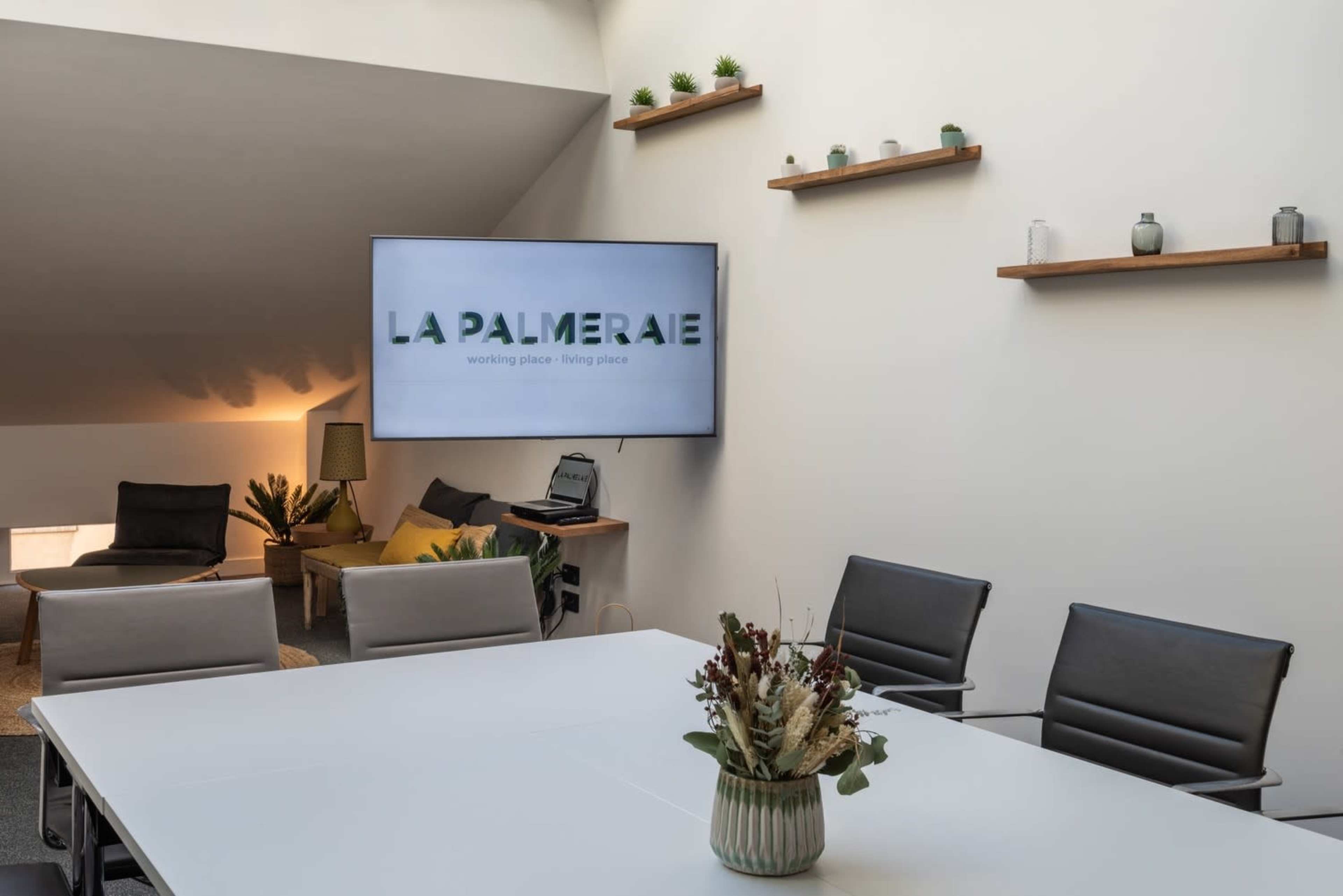 A modern meeting room features a large white table surrounded by black chairs, with a screen displaying the text "LA PALMERAIE" and several decorative shelves along the wall.
