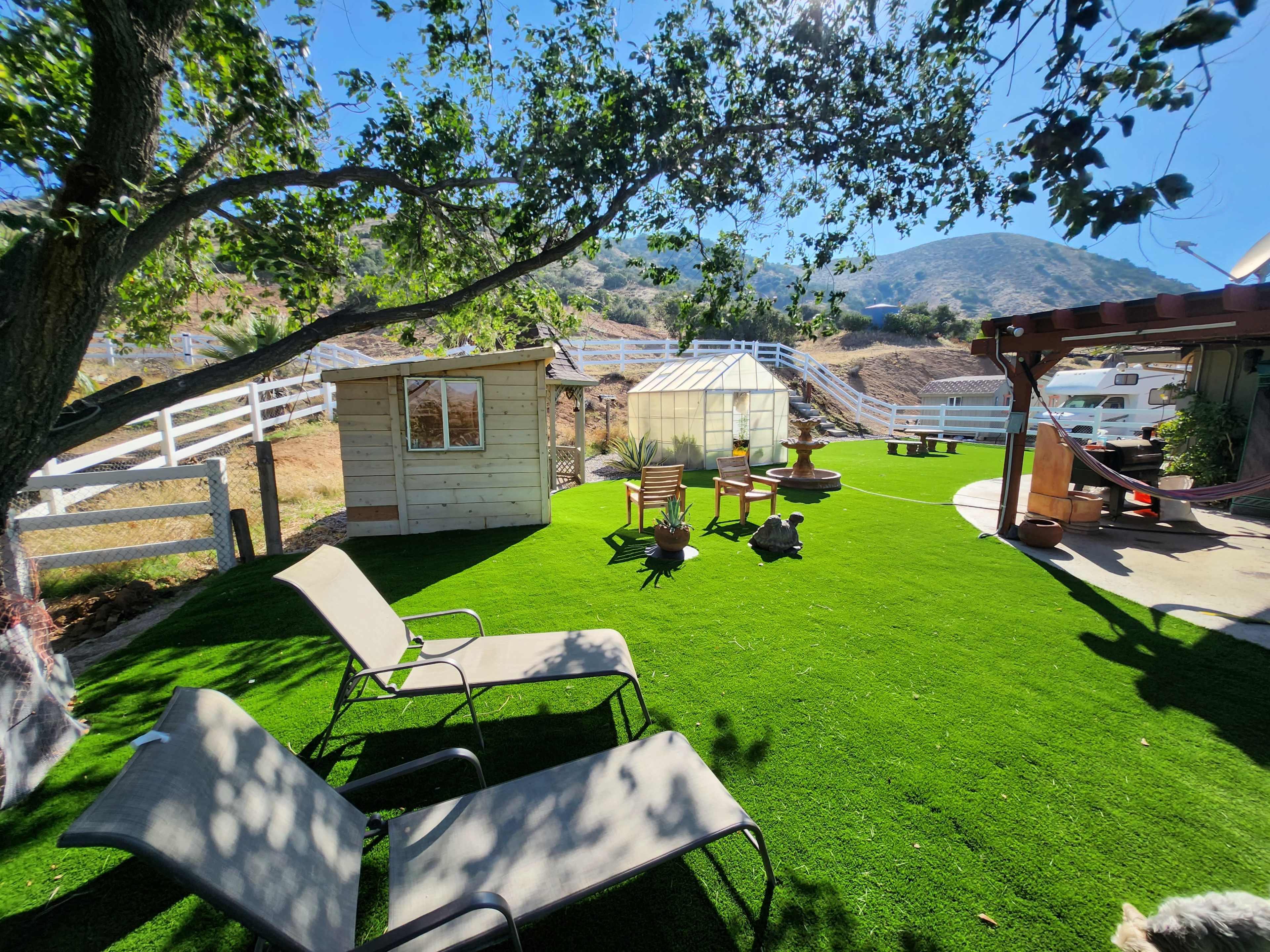 A green, grassy yard with two lounge chairs, a small shed, a greenhouse, and seating arranged around a fire pit, all set against a backdrop of hills and a blue sky.