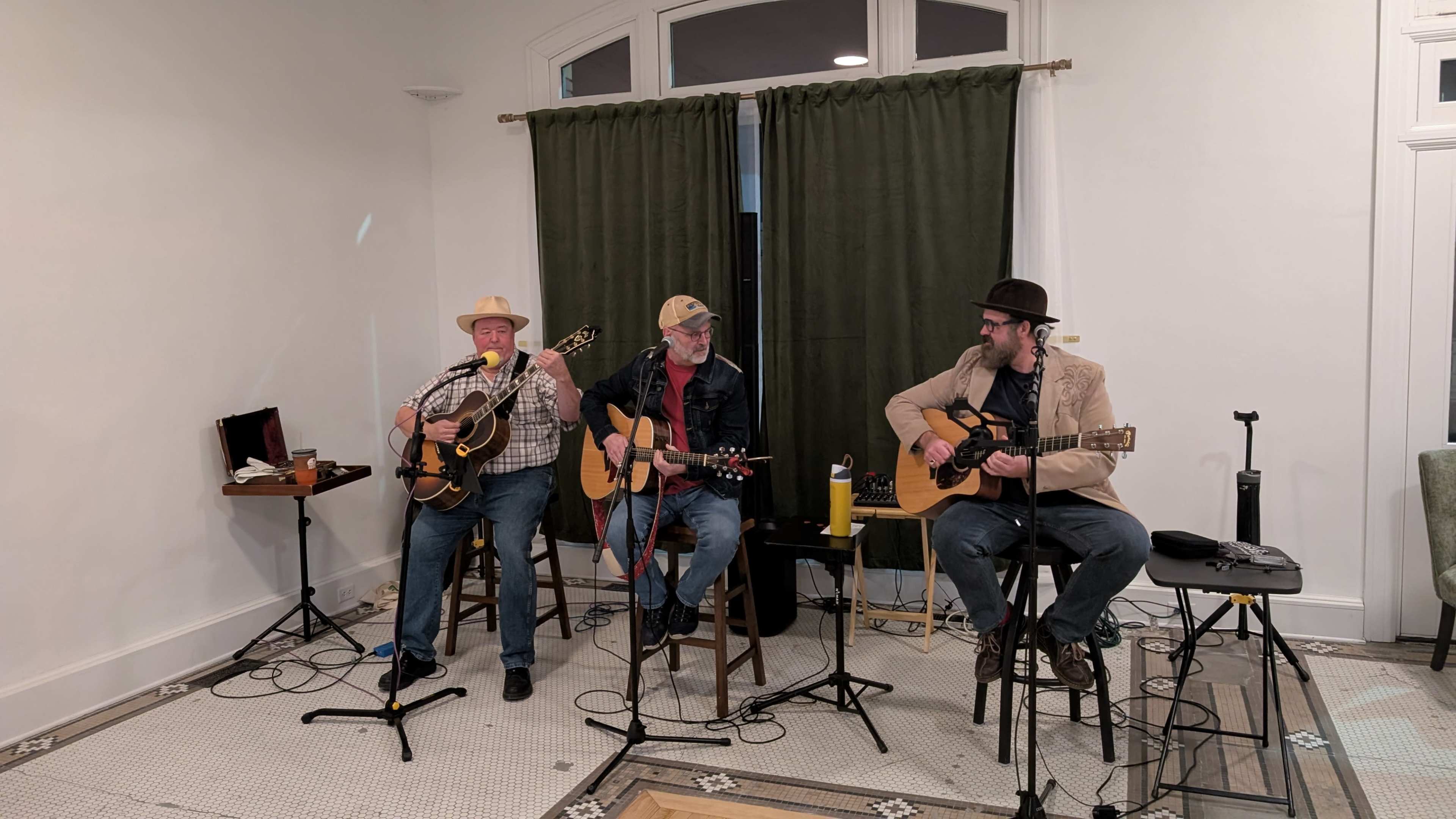 Three musicians play acoustic guitars while seated on stools in a room with green curtains.
