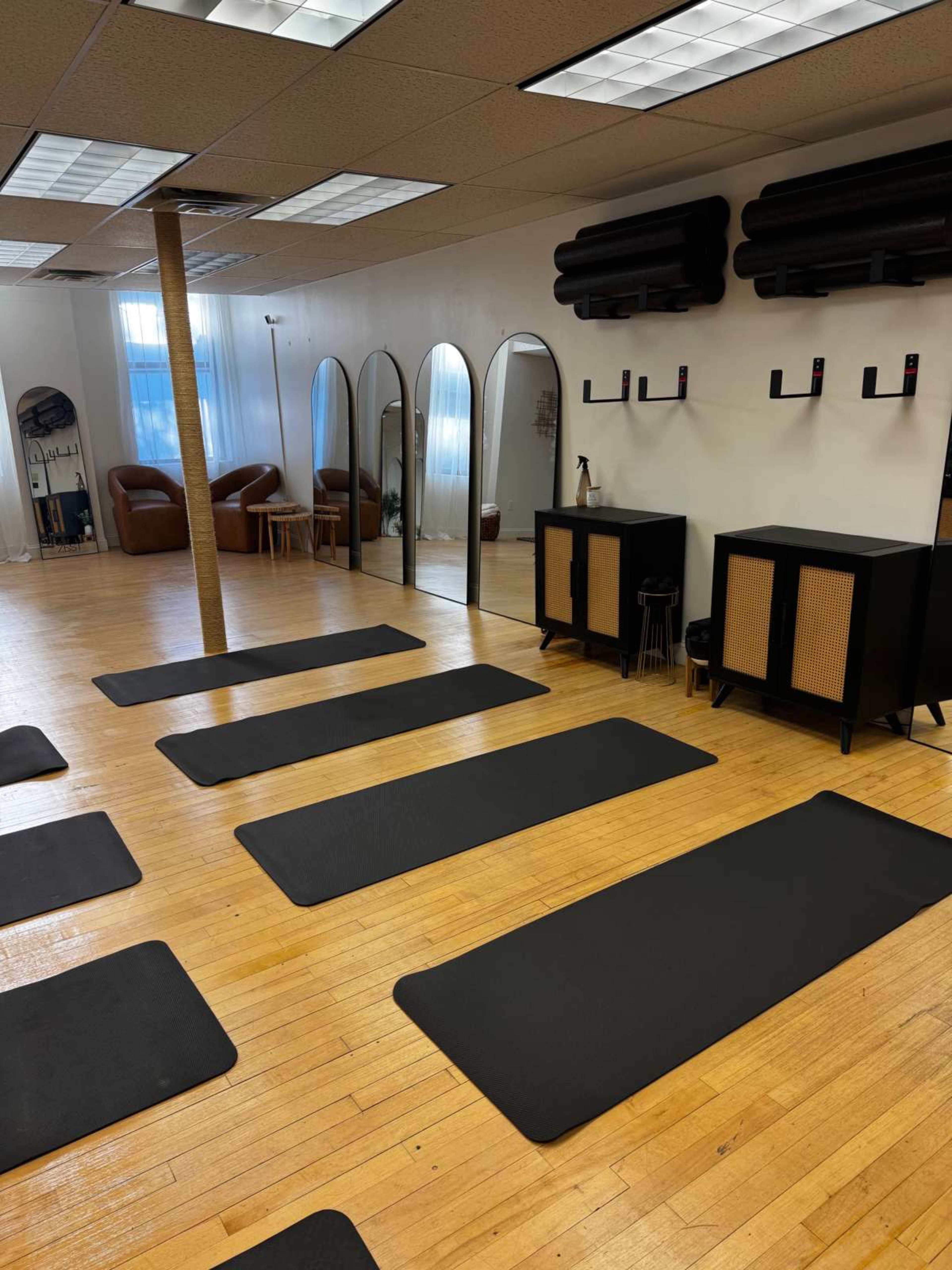 The image shows a fitness studio with multiple exercise mats arranged on a wooden floor, mirrors on one wall, and seating in the background.