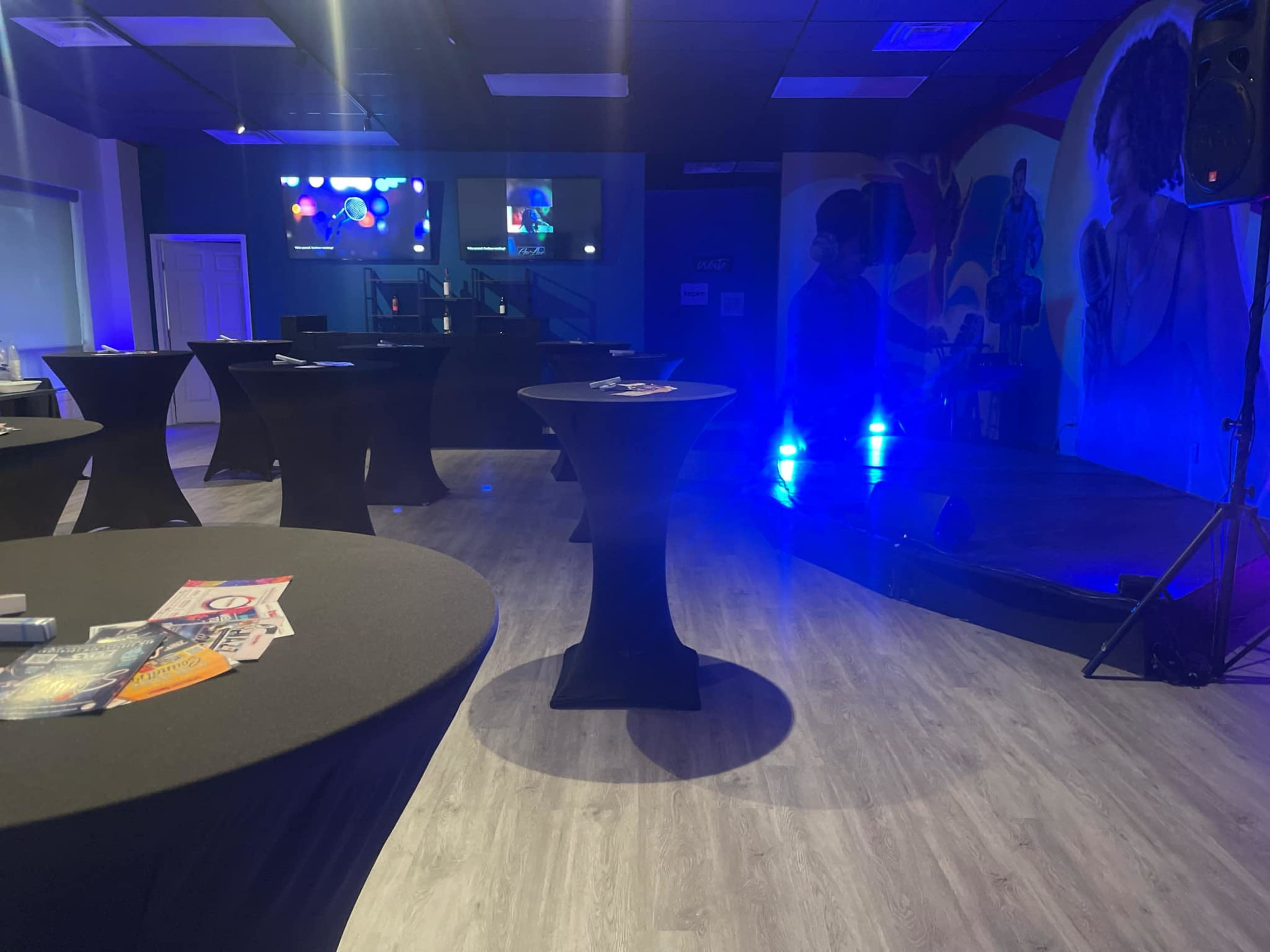 The image shows an indoor event space with black cocktail tables, a small stage, and colorful wall murals, all under blue lighting.