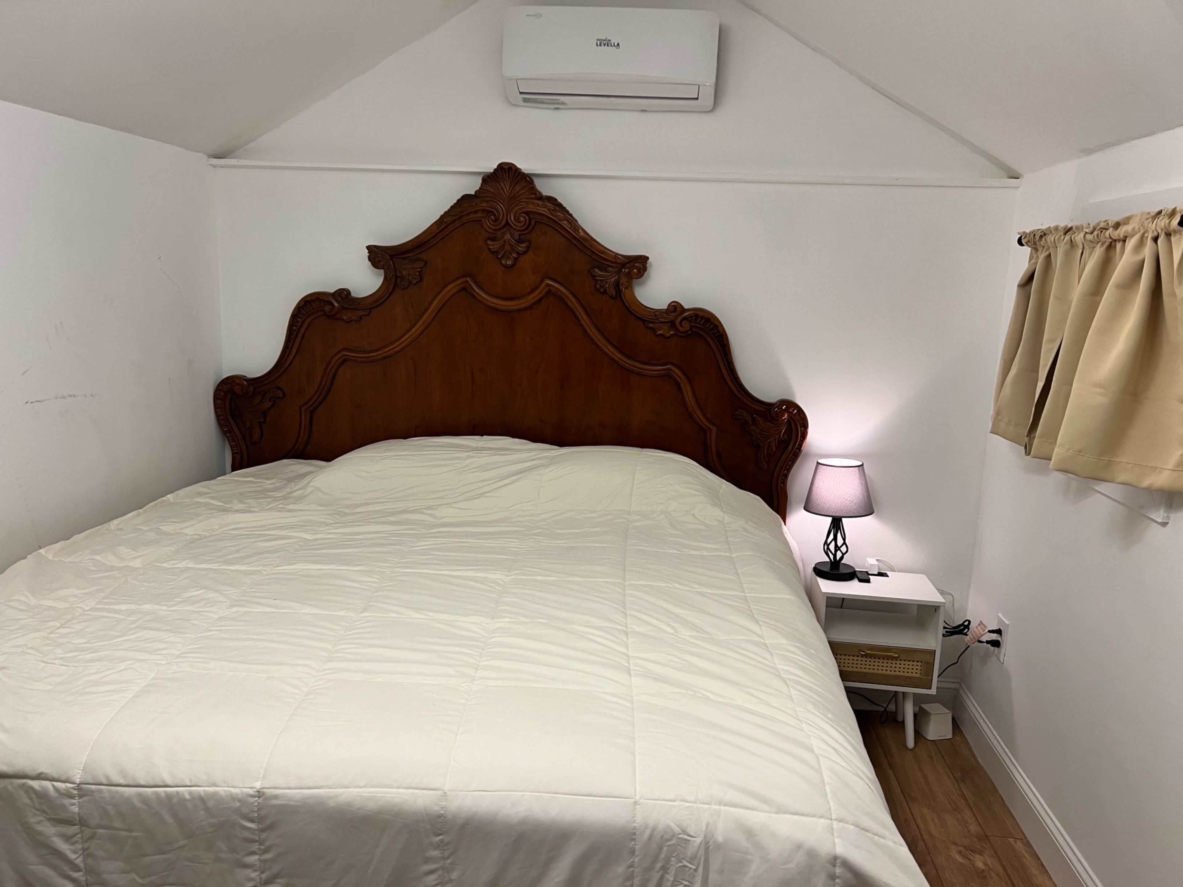A room with a wooden bed frame, a white comforter, and a nightstand with a lamp beside the bed.
