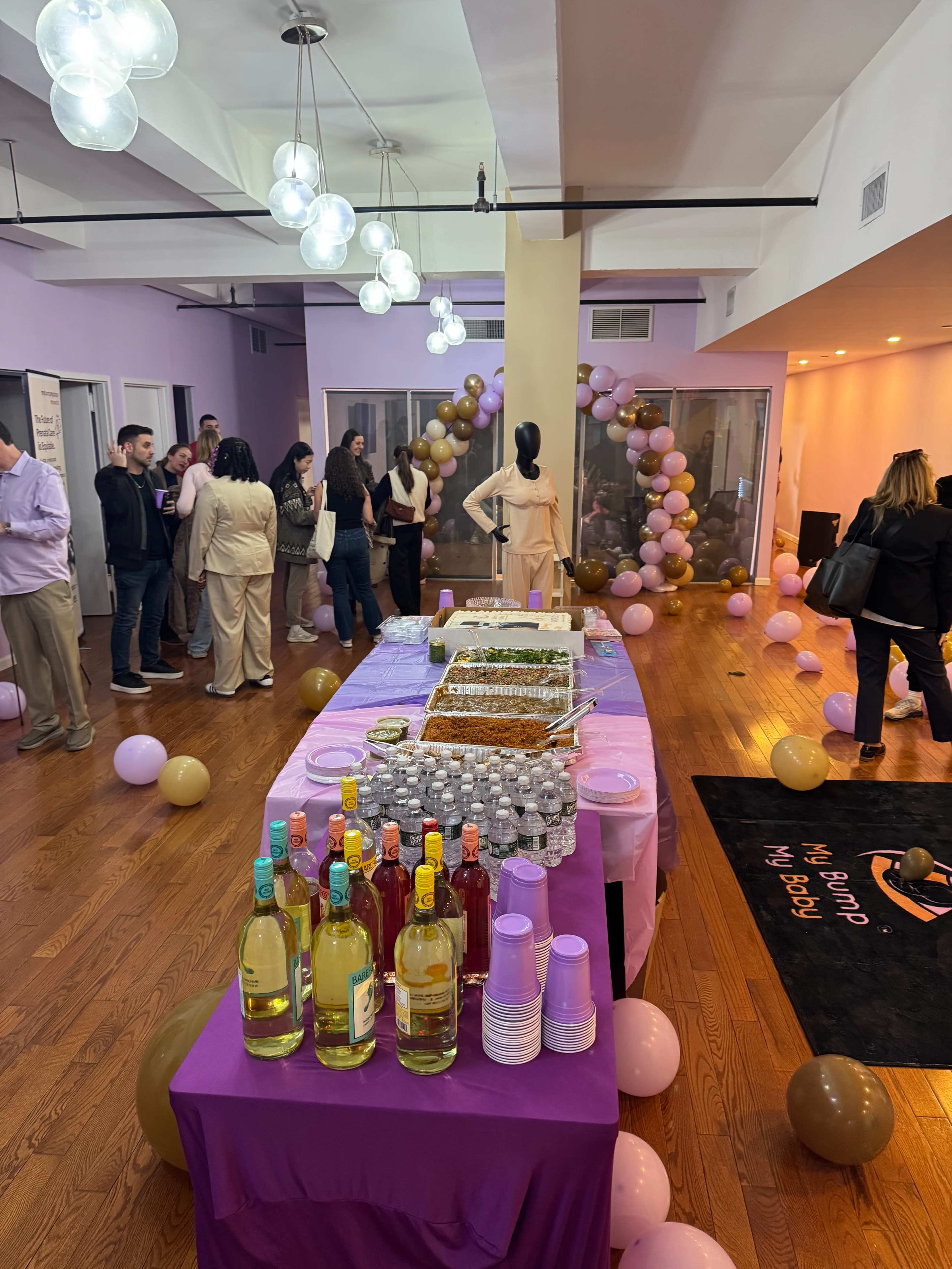 A party is taking place in a spacious room decorated with balloons, where guests mingle and a table is set with food and drinks.