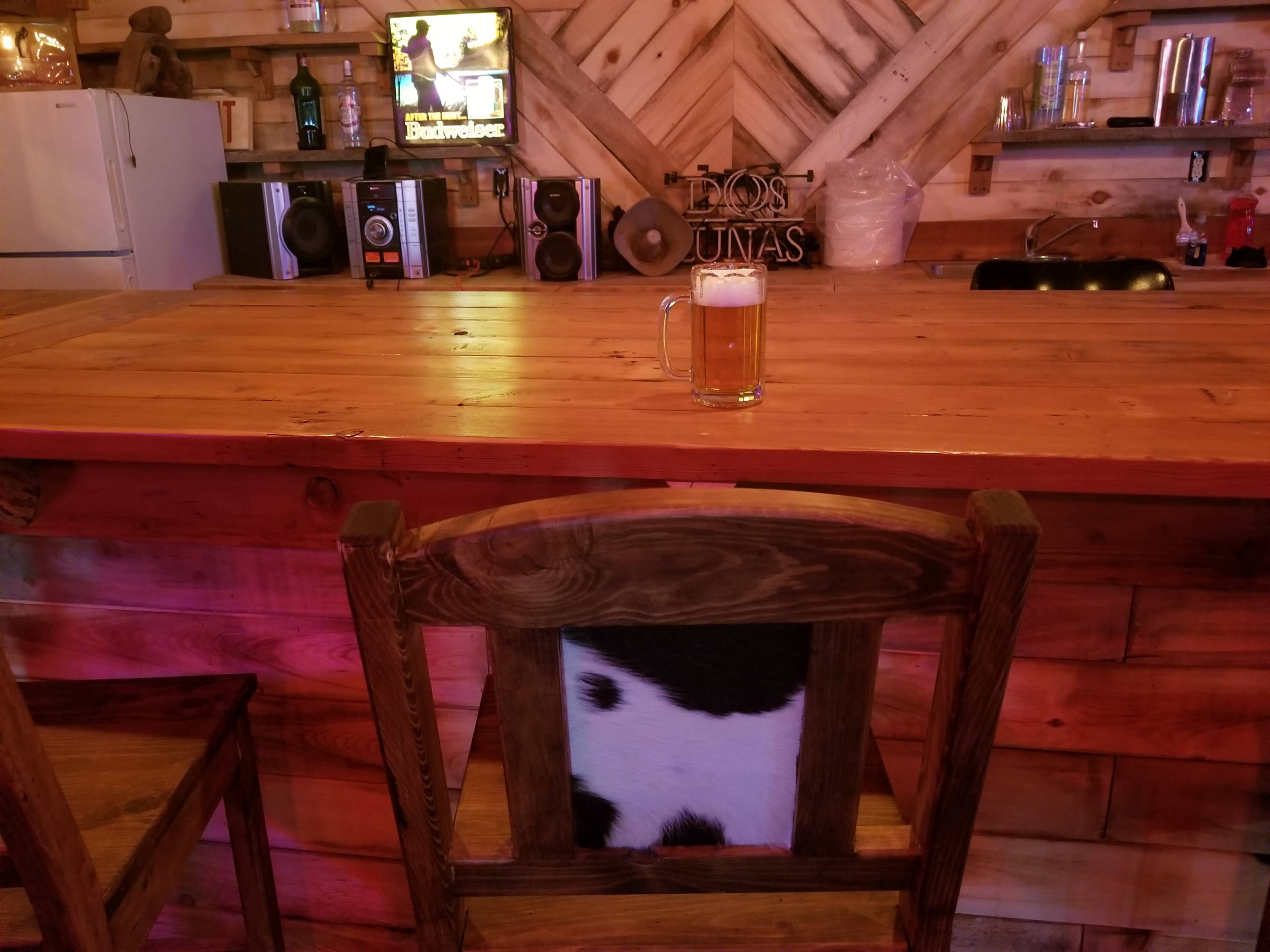 A wooden bar counter is set with a glass of beer in front of an empty chair, against a backdrop of rustic decor and bar equipment.