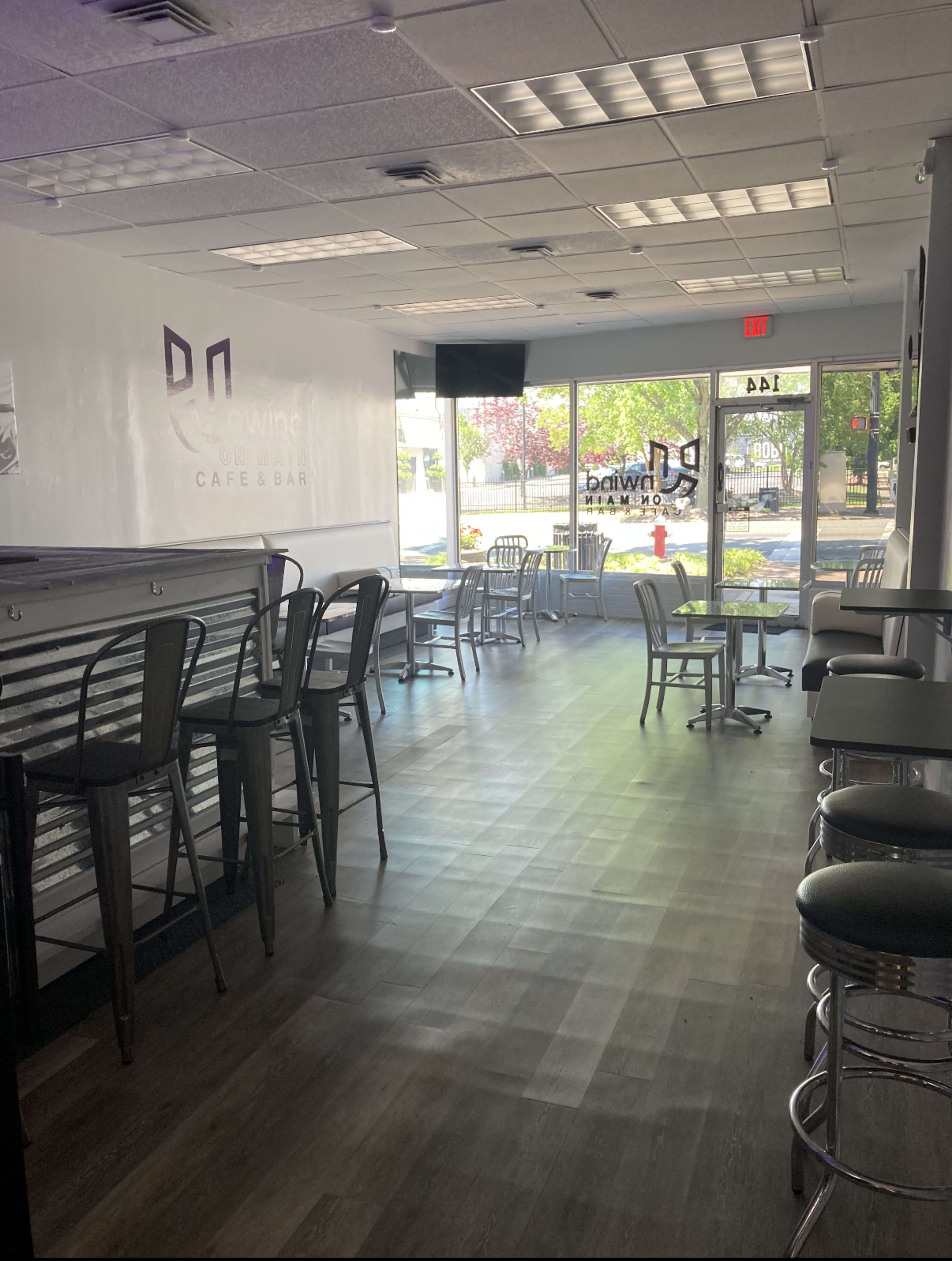 The image shows the interior of a cafe bar with a row of empty chairs and tables, illuminated by natural light from large windows.
