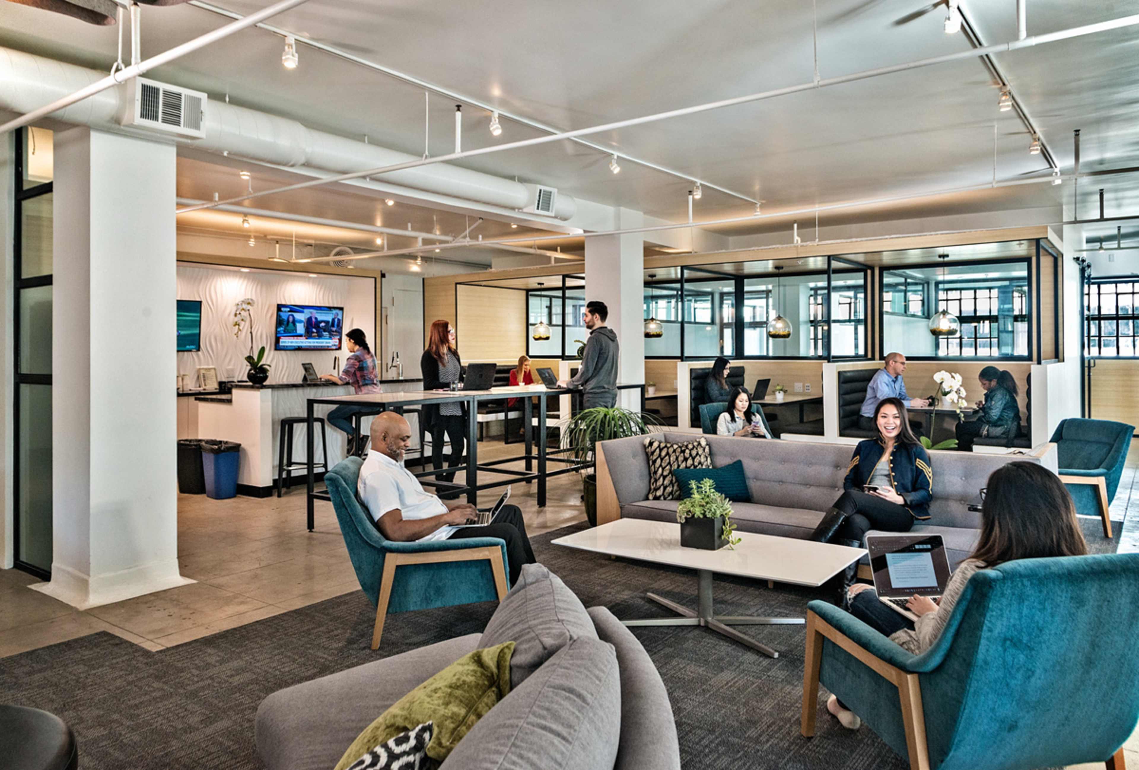 The image shows a modern office space with various groups of people engaging in different activities, including conversations, working on laptops, and standing at a bar-like counter.