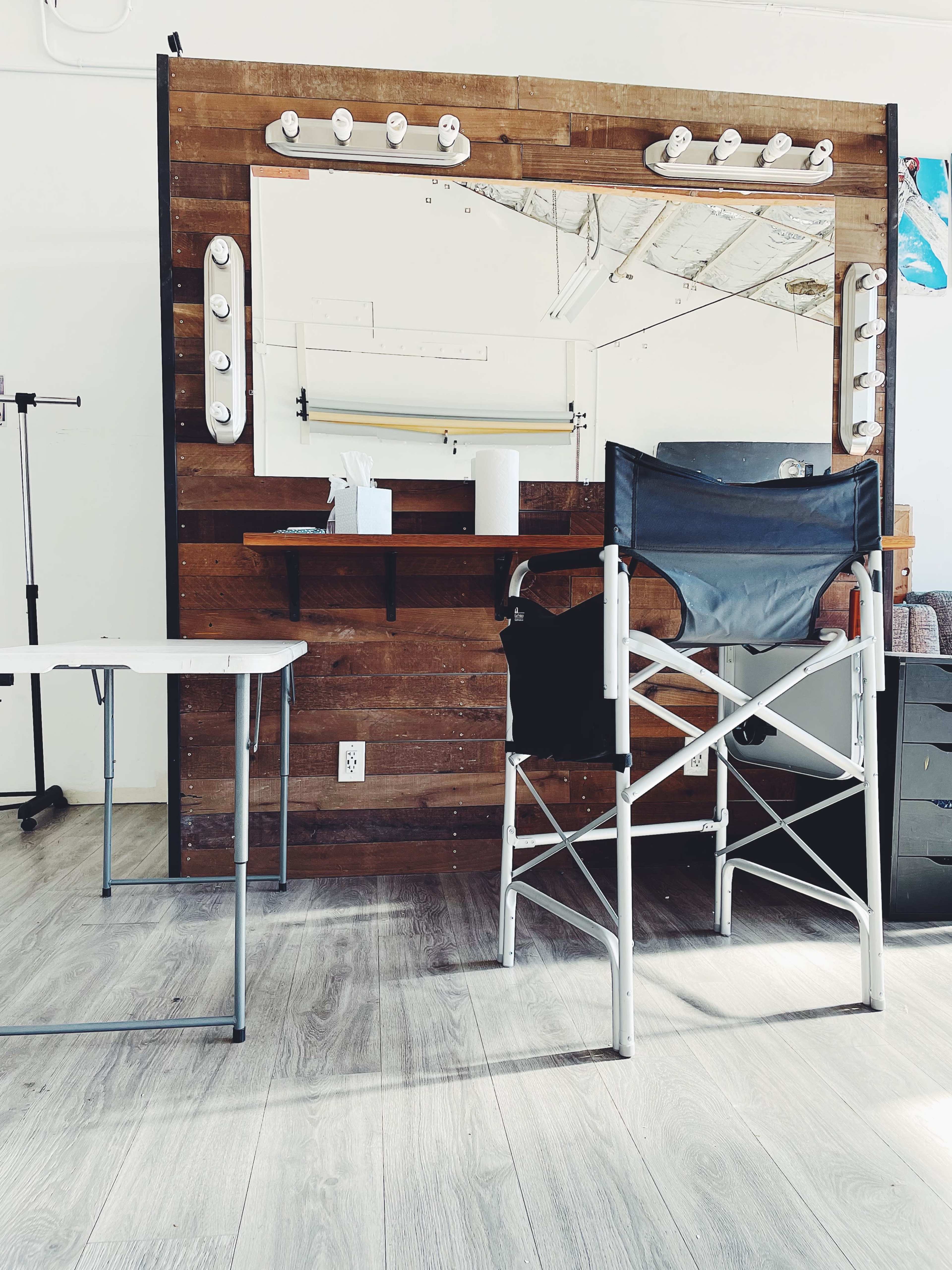 A wooden vanity mirror with bright bulbs, a folding chair, and a small table set in a bright, spacious room.