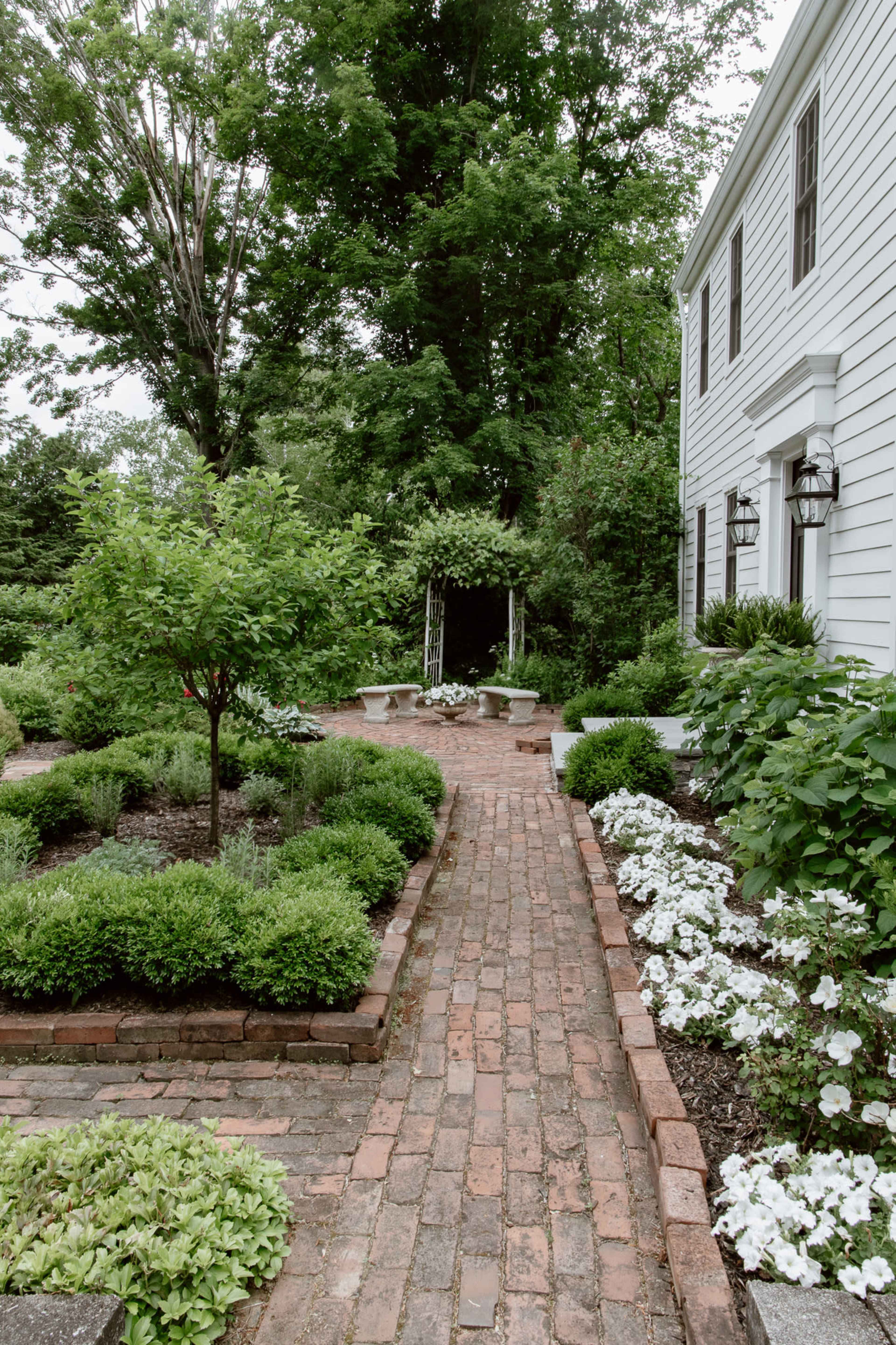 A brick pathway lined with greenery and flower beds leads to a small arbor in a garden next to a white house.