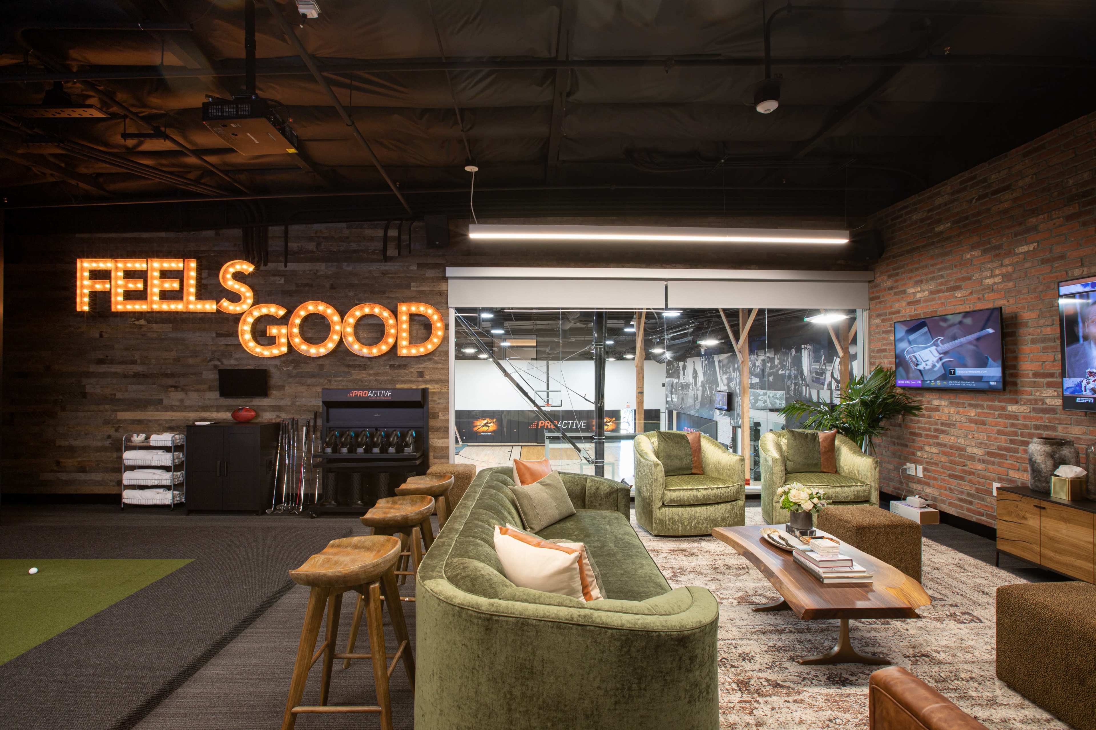 The image shows a modern, industrial-style lounge area featuring a large "FEELS GOOD" neon sign, comfortable seating, and an open view to a gym space.