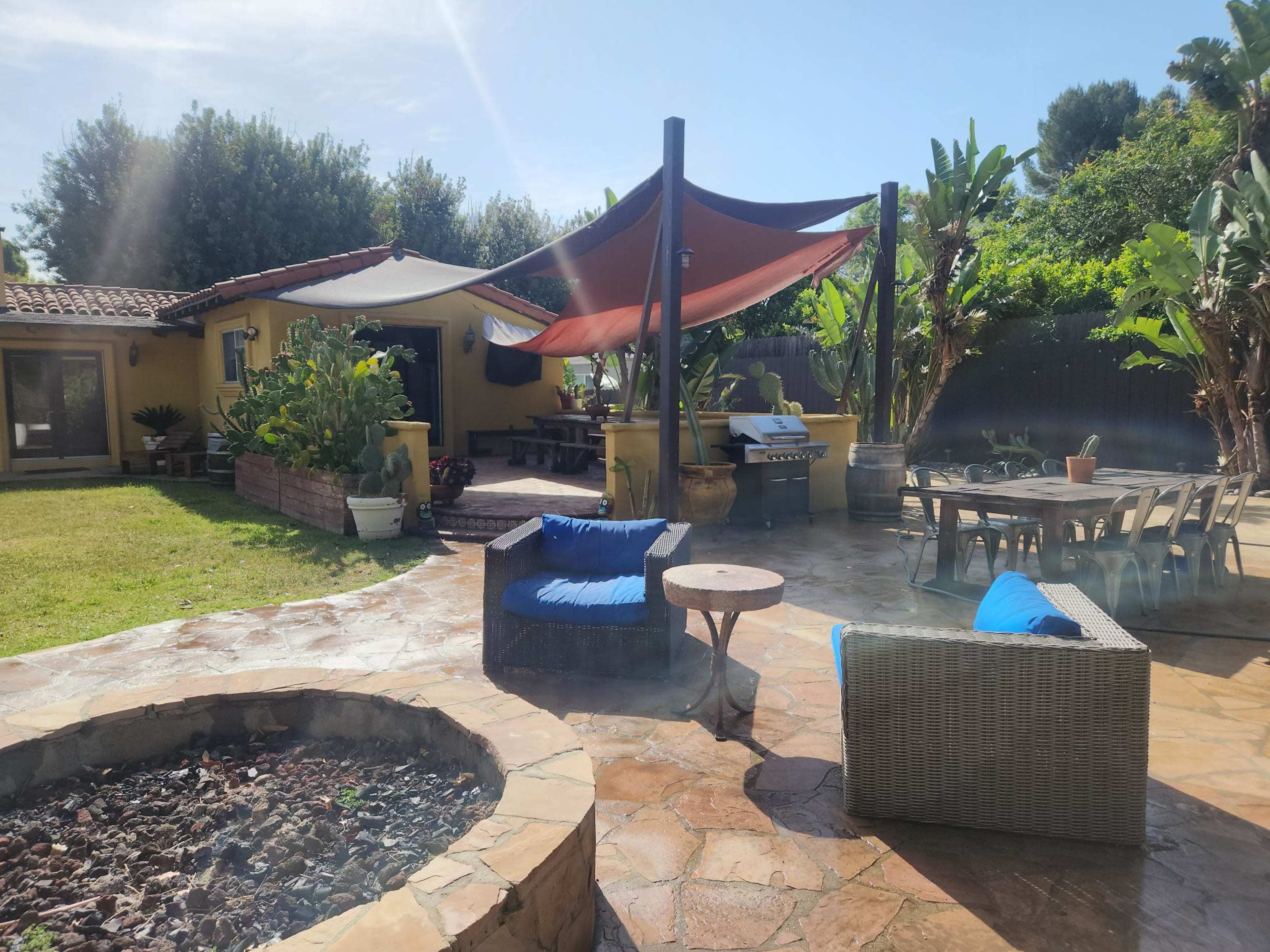 The image shows a patio area with outdoor seating, a fire pit, and a grill, surrounded by lush greenery and a sunny sky.