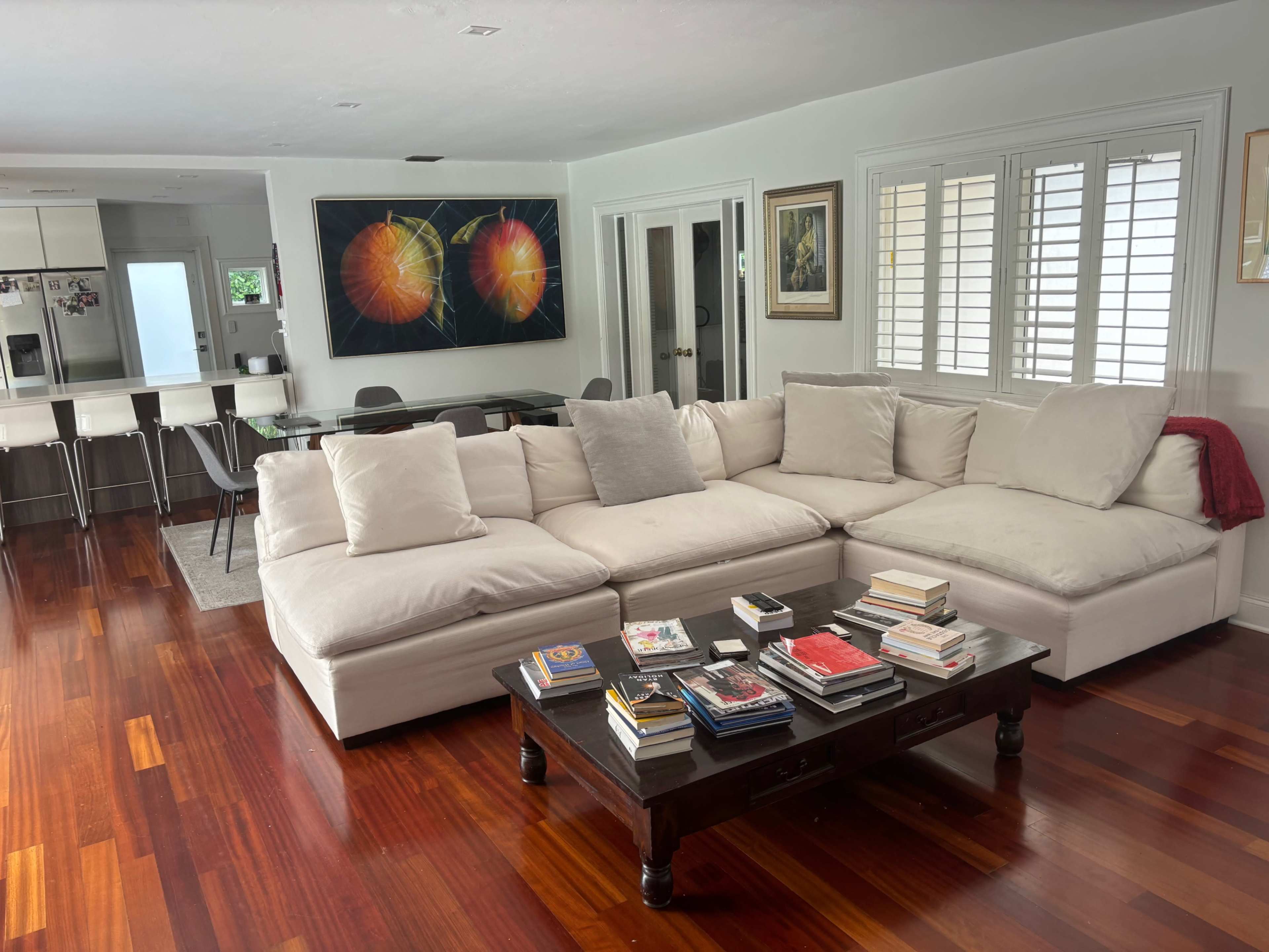 A sectional sofa with white cushions is arranged in a living room next to a coffee table stacked with books, with a dining area visible in the background.