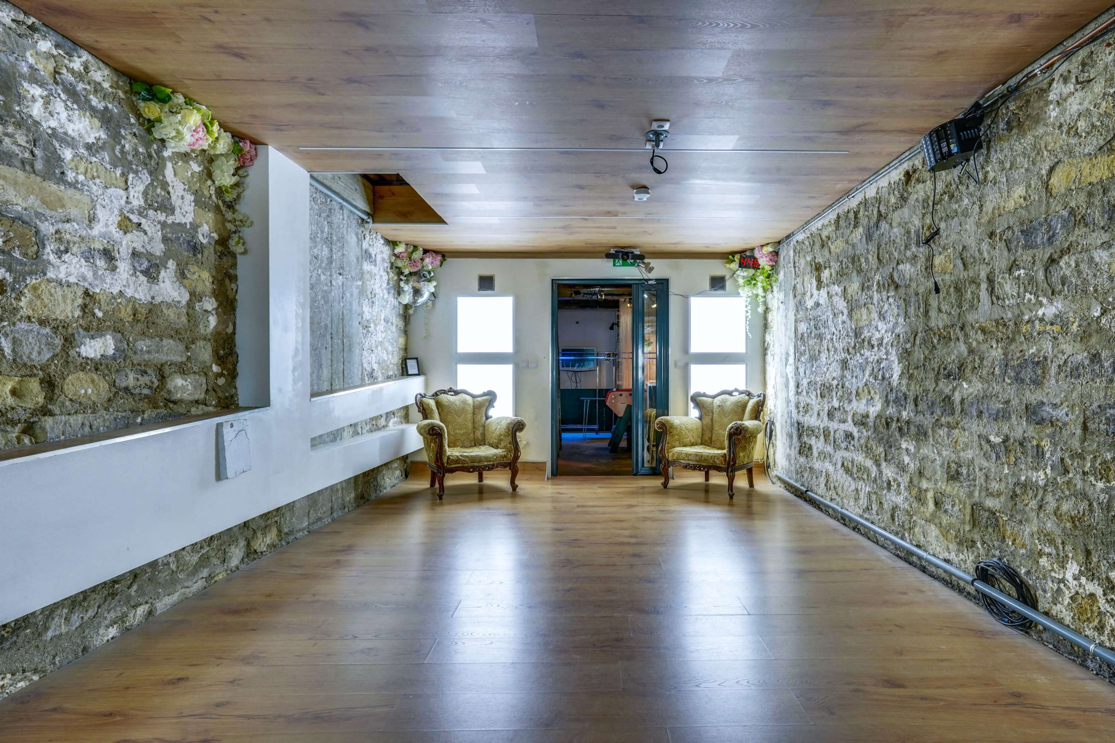 The image shows a room with stone walls and wooden flooring, featuring two upholstered chairs and a doorway at the back.