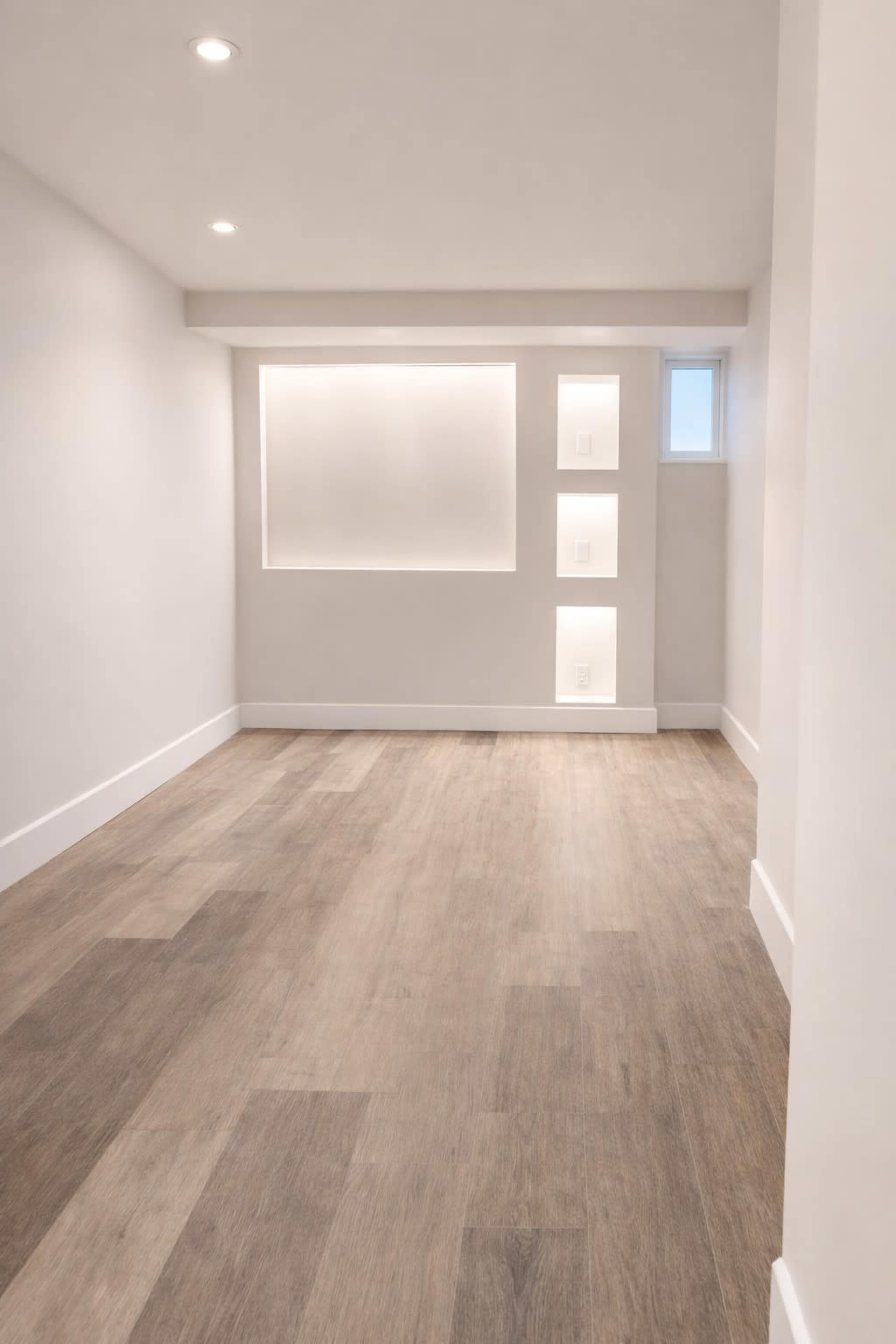 The image shows a sparsely furnished, modern room with light-colored walls, wooden flooring, and built-in light fixtures on one side.