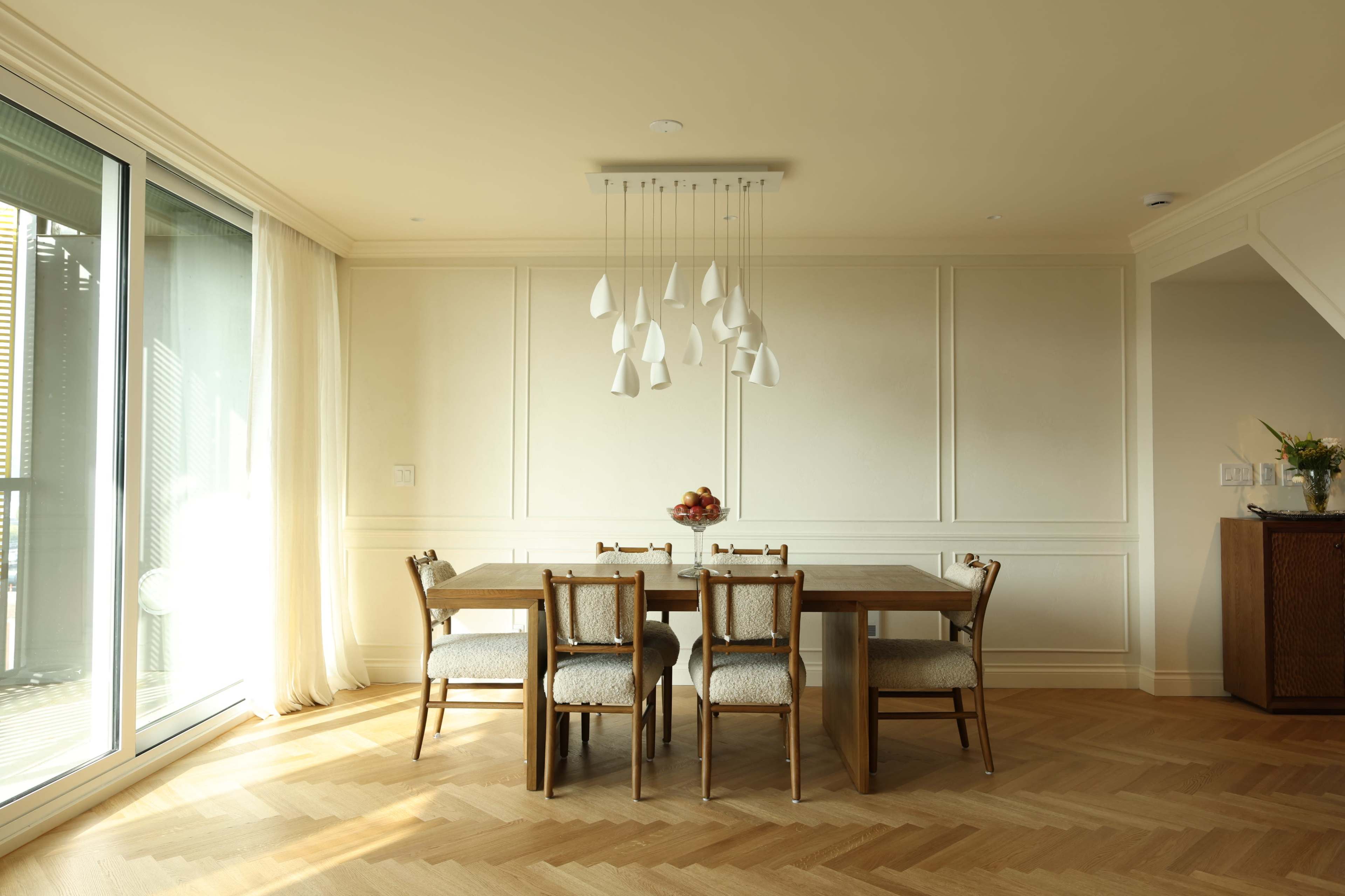 A dining area features a wooden table surrounded by four chairs and is illuminated by pendant lights, with large glass doors leading to an outside space.