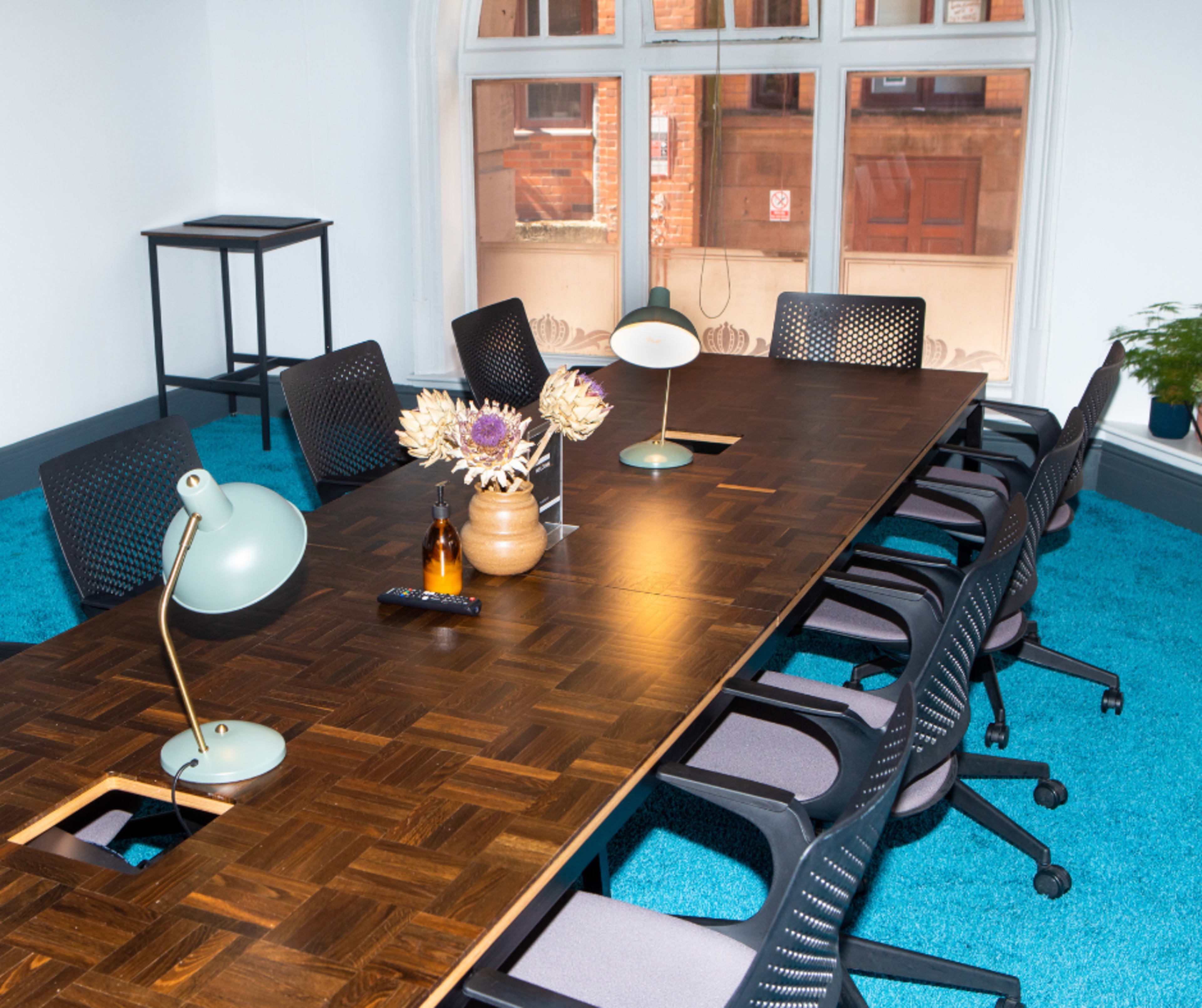 A long wooden conference table is surrounded by black office chairs in a brightly lit room with blue carpet and a large window.