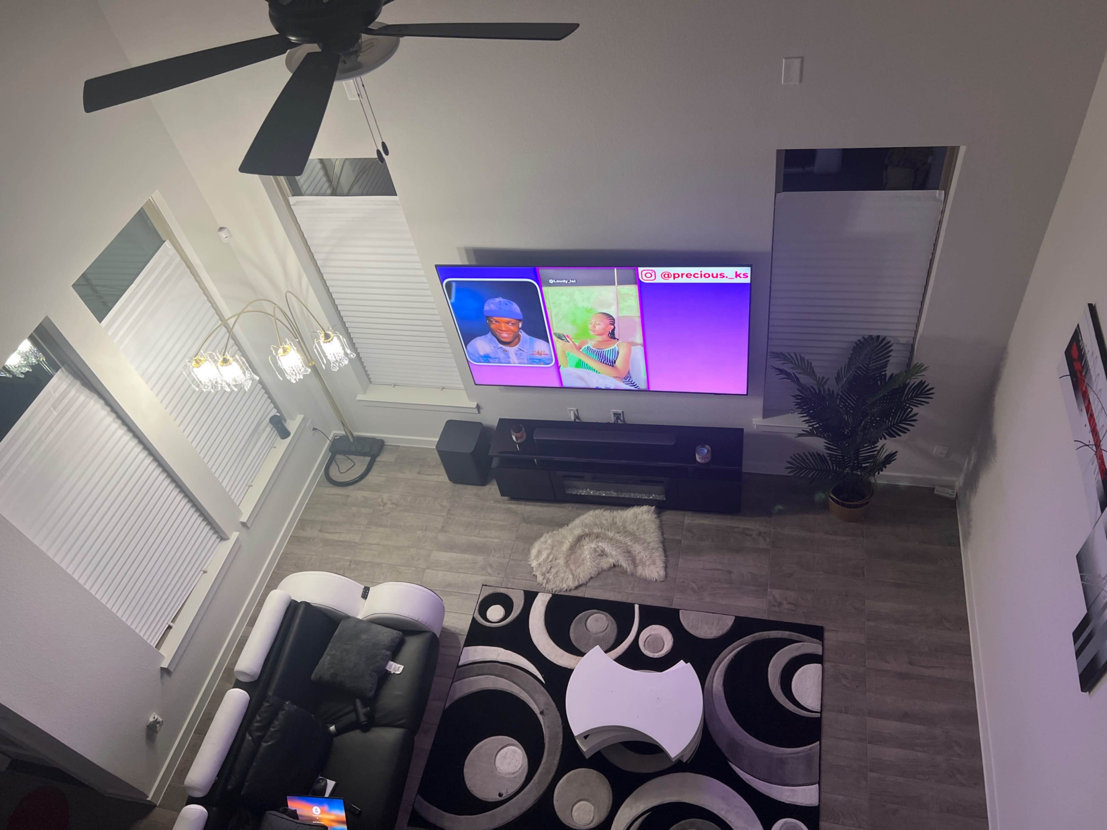 The image shows a living room from an overhead view, featuring a large television displaying two images, a black and white circular rug on the floor, and modern furniture including a black couch and white coffee table.