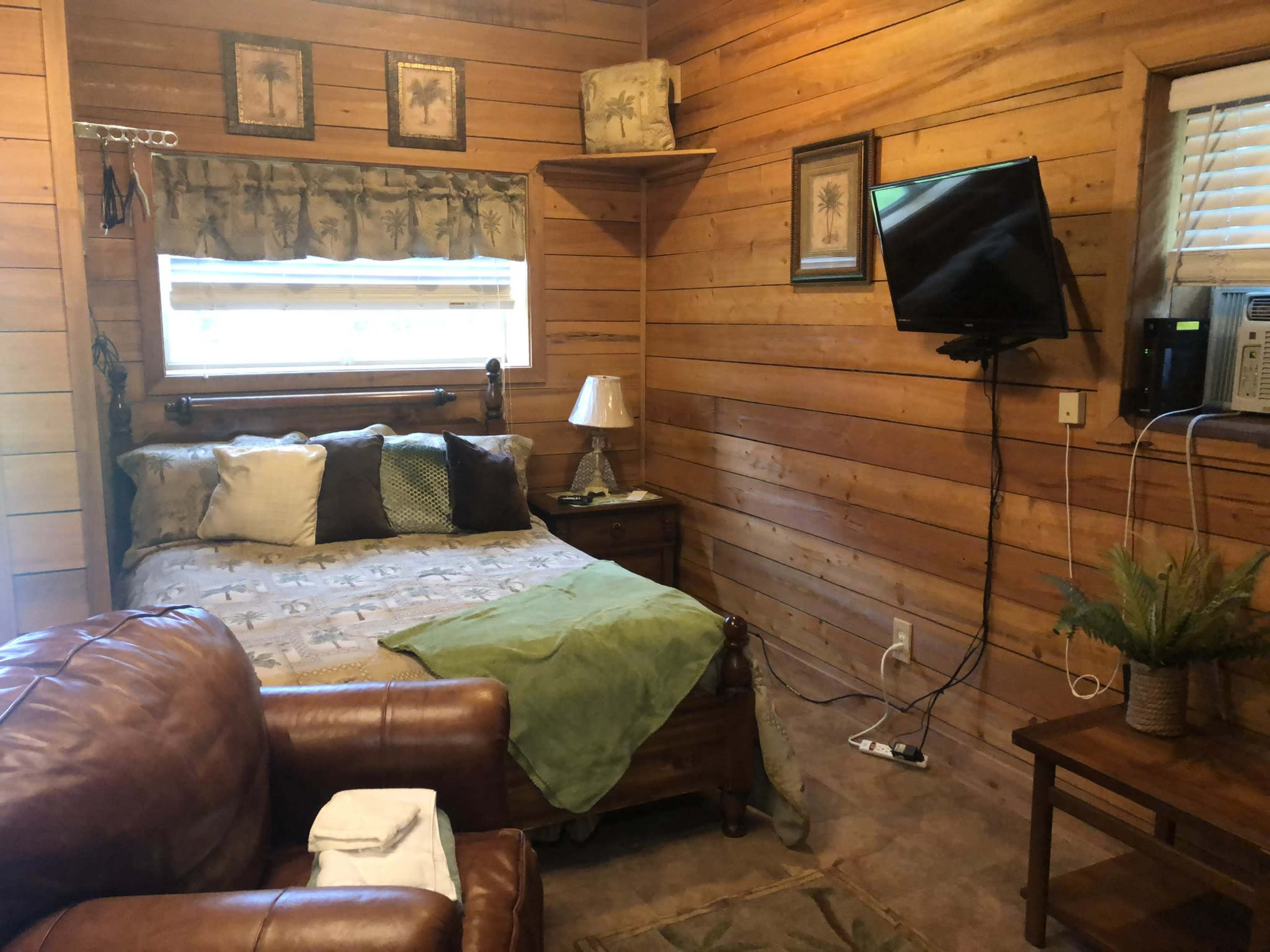 A cozy wooden room featuring a bed with various pillows, a small table with a lamp, a television mounted on the wall, and a comfortable chair.