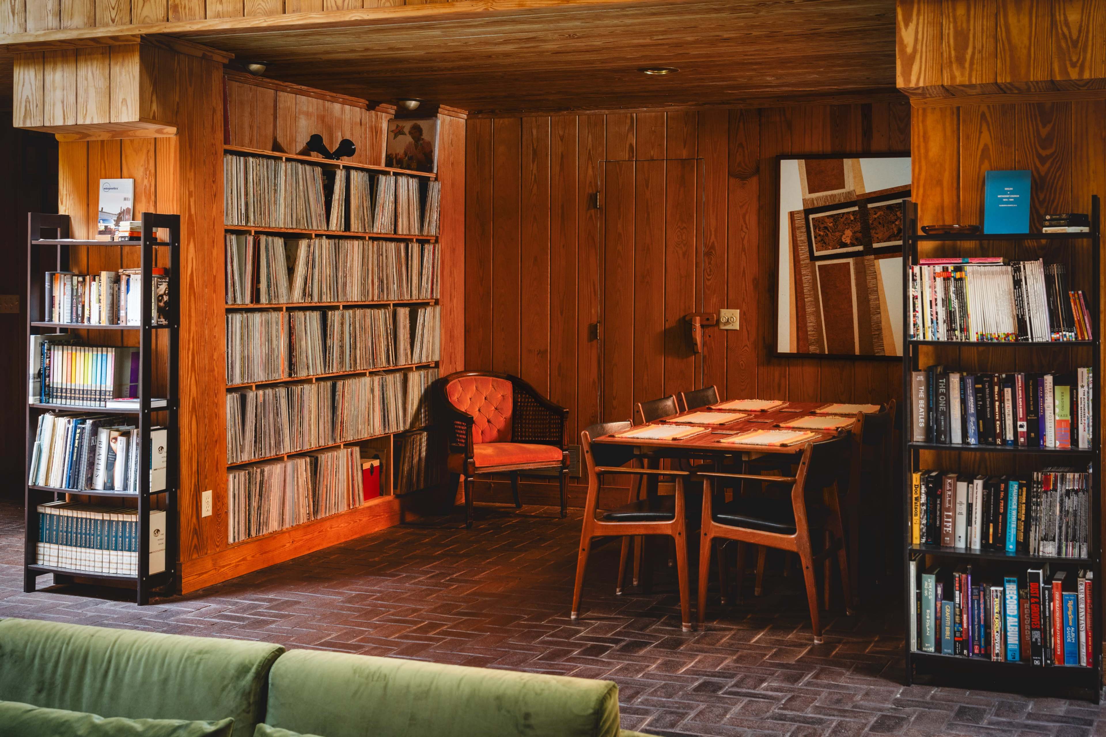 A cozy room features wooden walls lined with vinyl records and bookshelves, a table with chairs, and a vintage armchair.