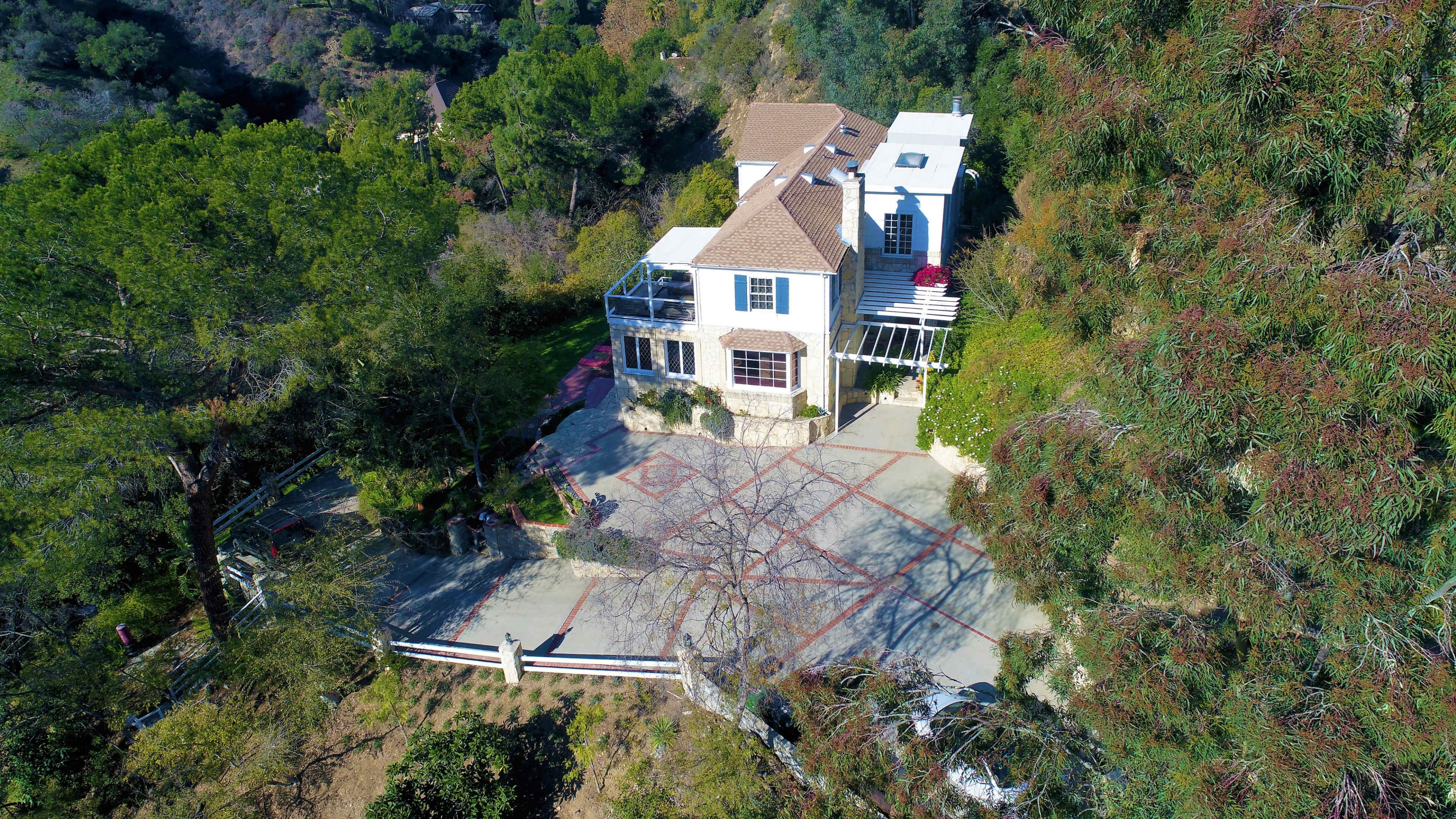 A large, two-story house situated on a hillside surrounded by greenery and a paved driveway.