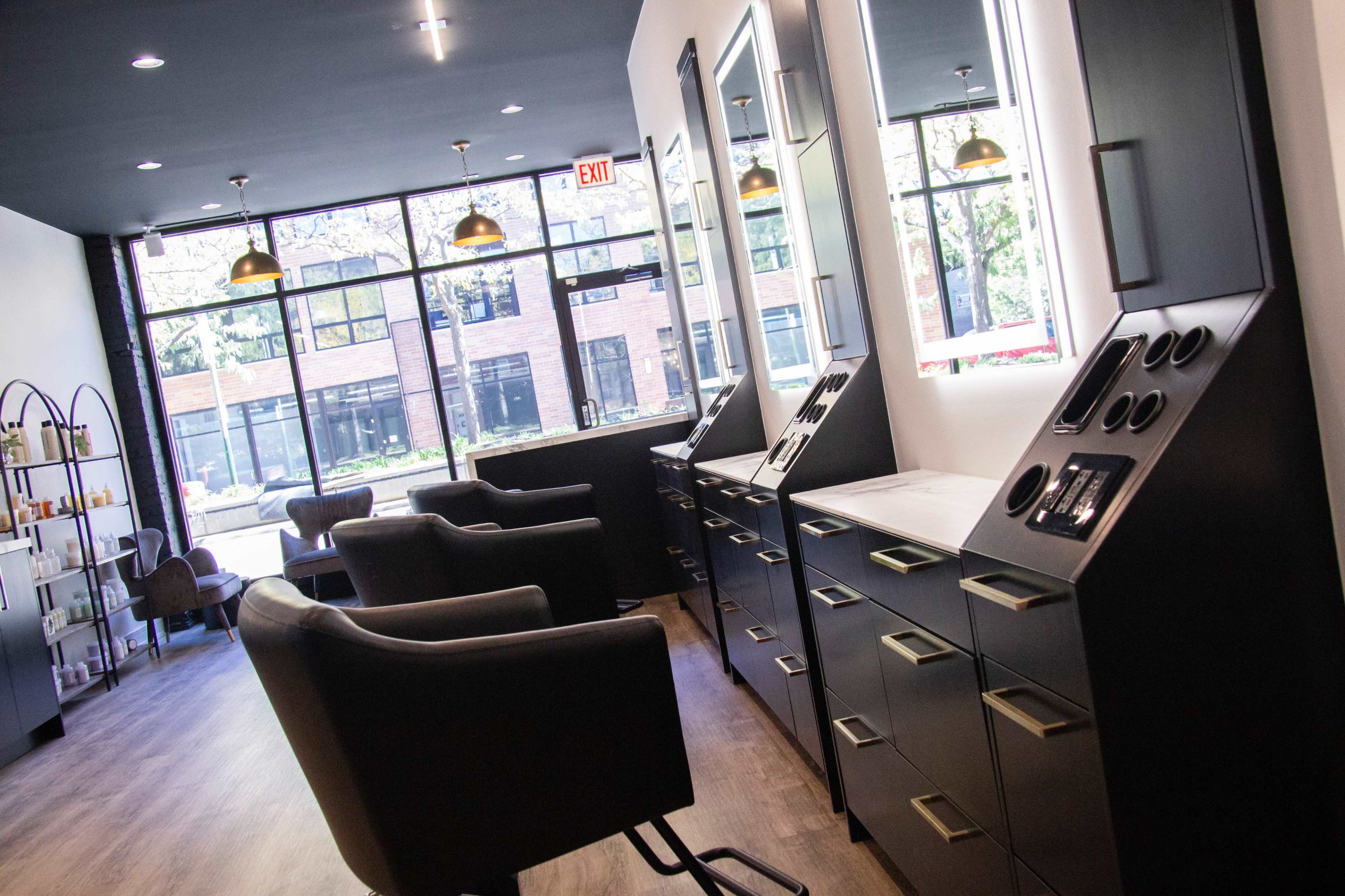The image shows a modern salon interior with black chairs, vanity stations featuring mirrors and makeup products, and large windows allowing natural light to enter.