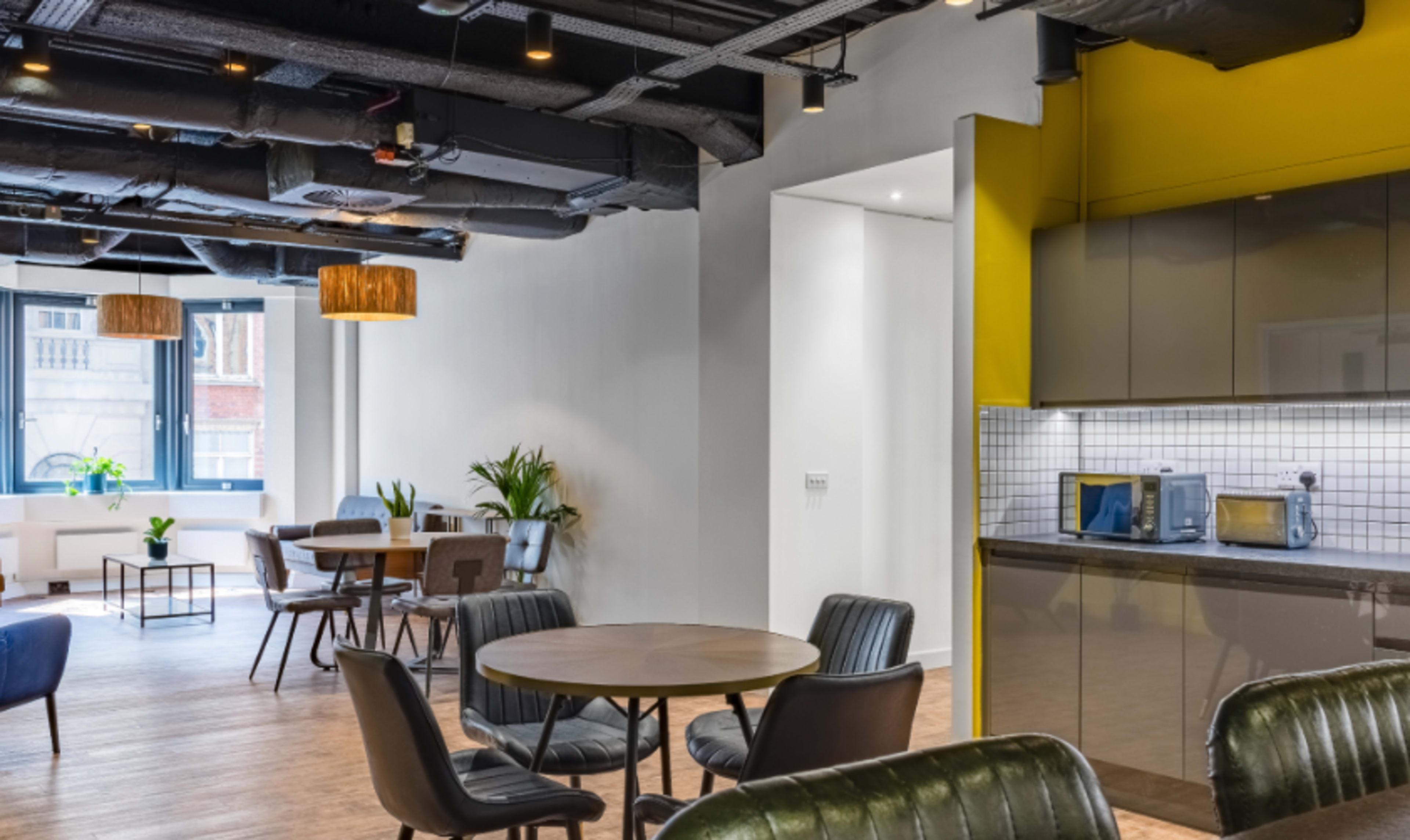 A modern office common area featuring round tables, black leather chairs, and a kitchenette with yellow accent walls.