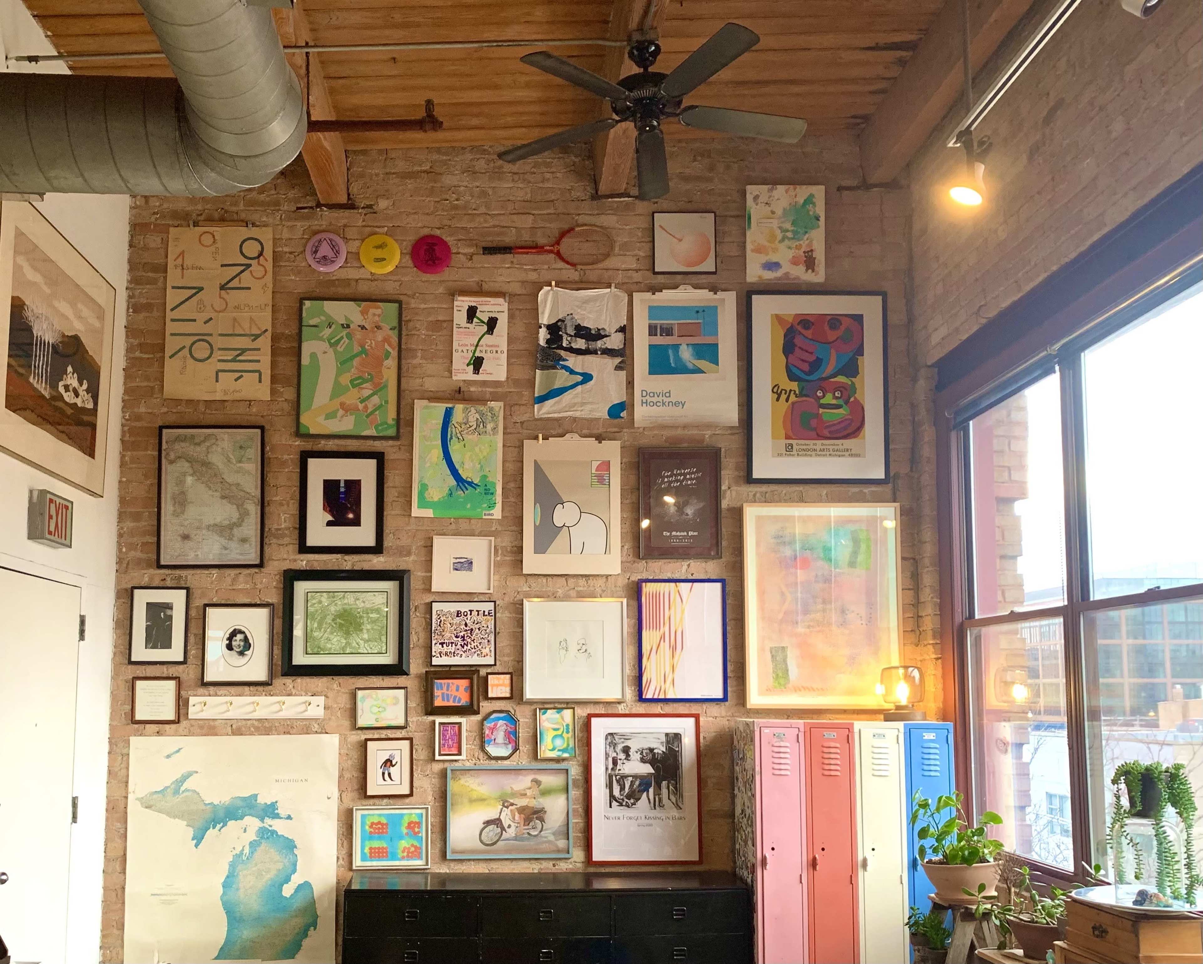 A wall covered with a variety of framed artworks and posters in a room with wooden beams and large windows.