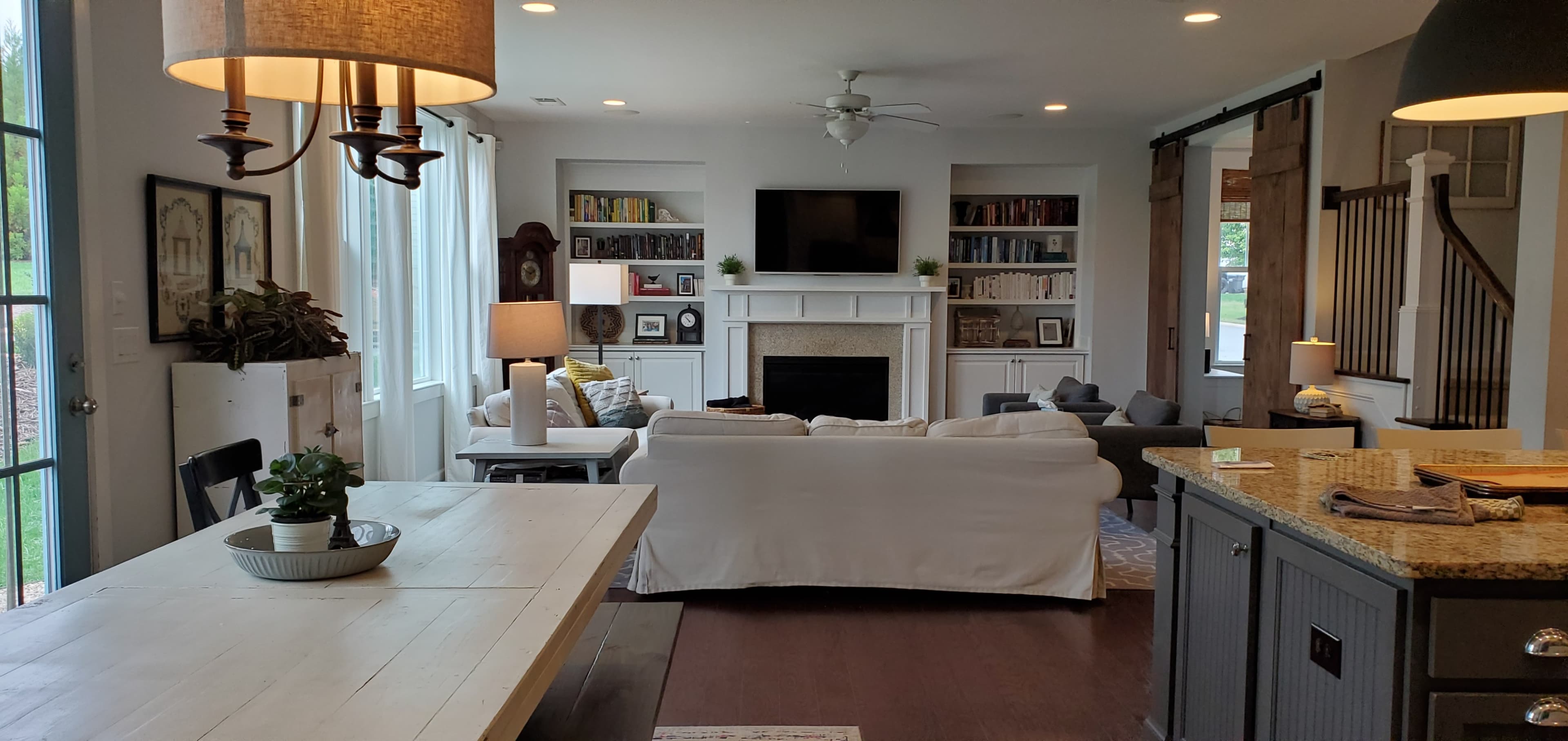 A light-filled living room features a cozy sofa, a fireplace with a TV above it, built-in bookshelves, and a dining area with a large table.